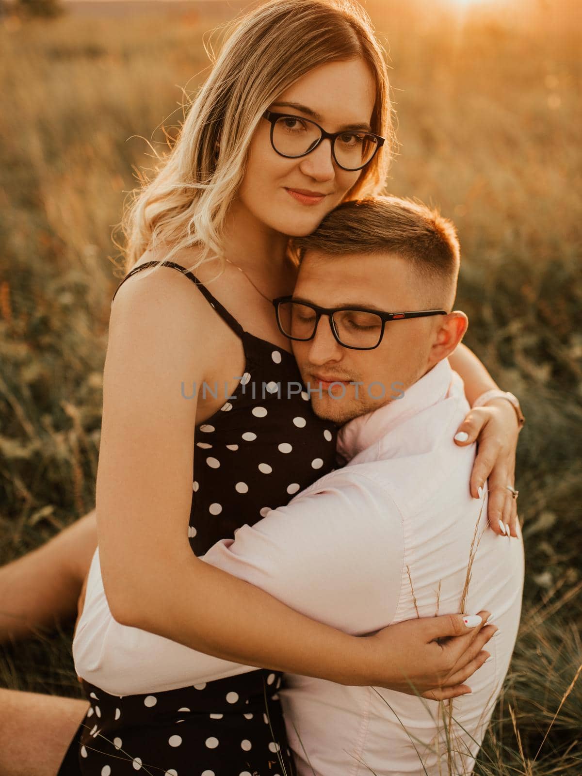 A girl in hat sits in hands guy on ground in middle of tall grass in meadow. couple of fair-haired fair-skinned people in love are resting in nature in field at sunset.