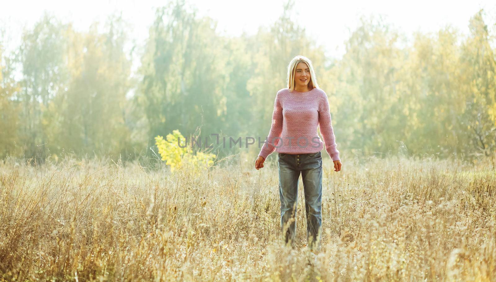 Charming blonde standing alone on the lawn among autumn or summer nature backlit by sunlight. Relaxation concept, Freedom concept.