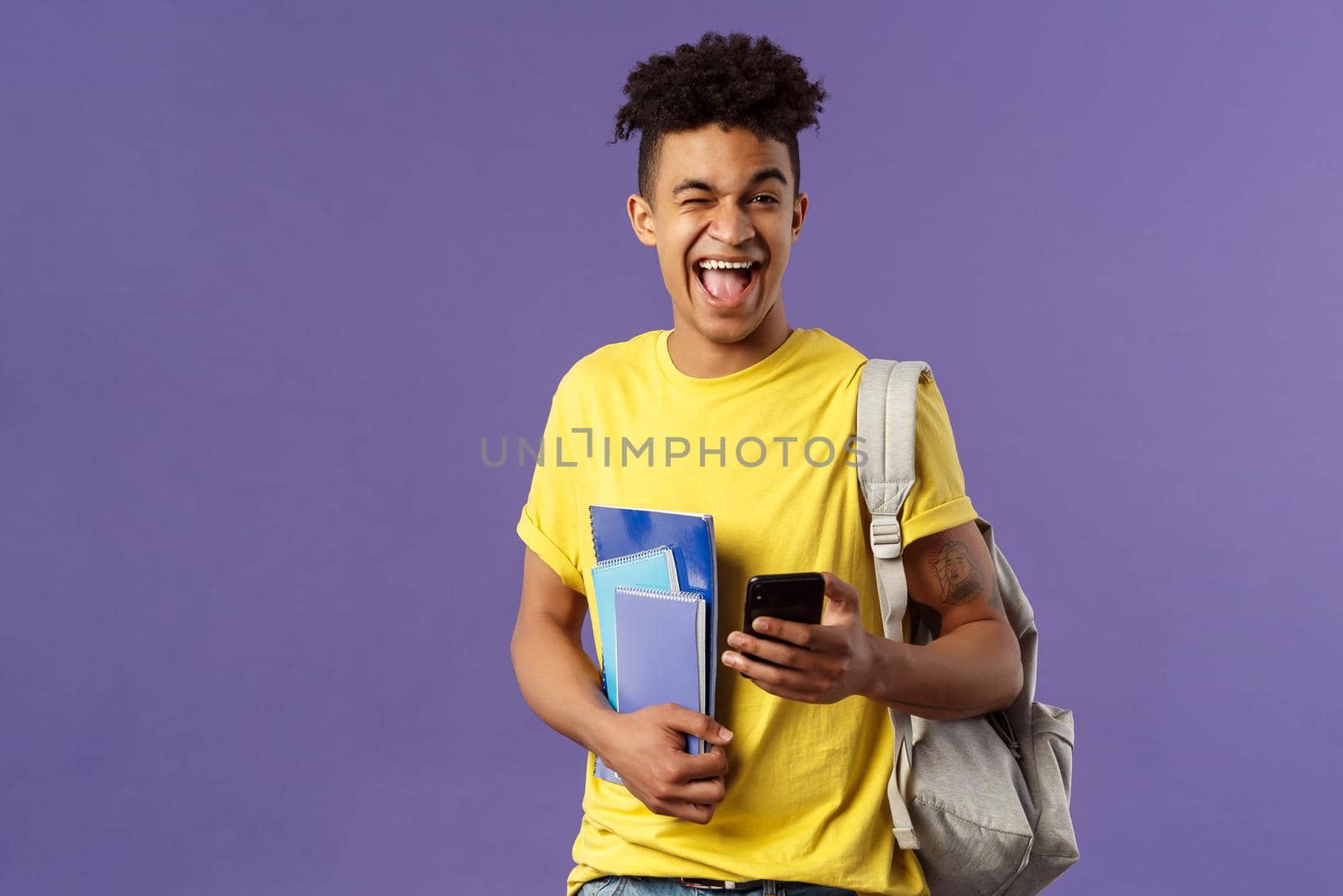 Back to school, university concept. Portrait of joyful, smiling happy man, student using mobile phone, wink at camera upbeat, holding backpack and notebooks, notes from lecture and smartphone by Benzoix