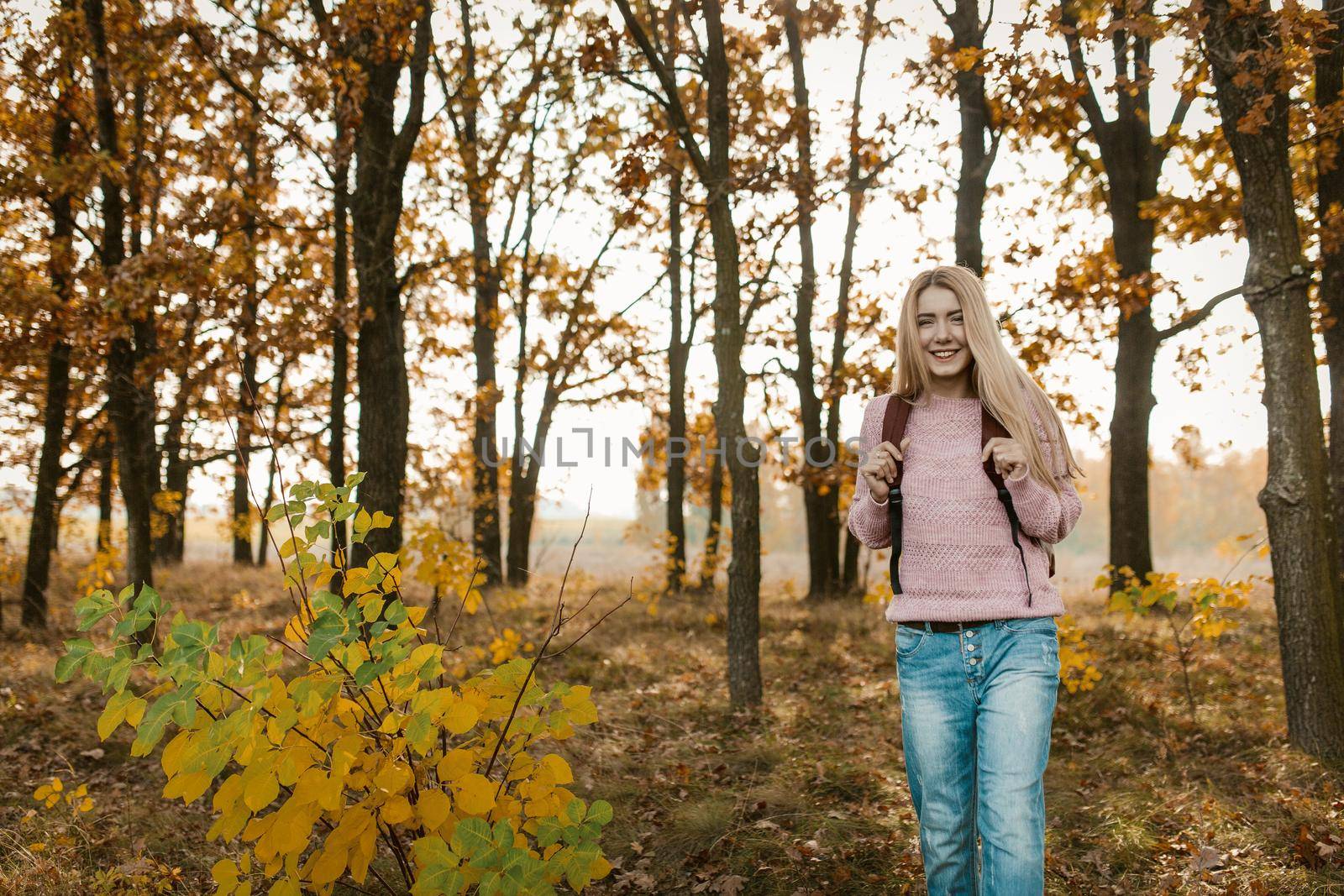 Traveling girl with a backpack smiles standing on beautiful autumn forest background. Caucasian blonde travels on nature on a sunny day. Hiking concept.