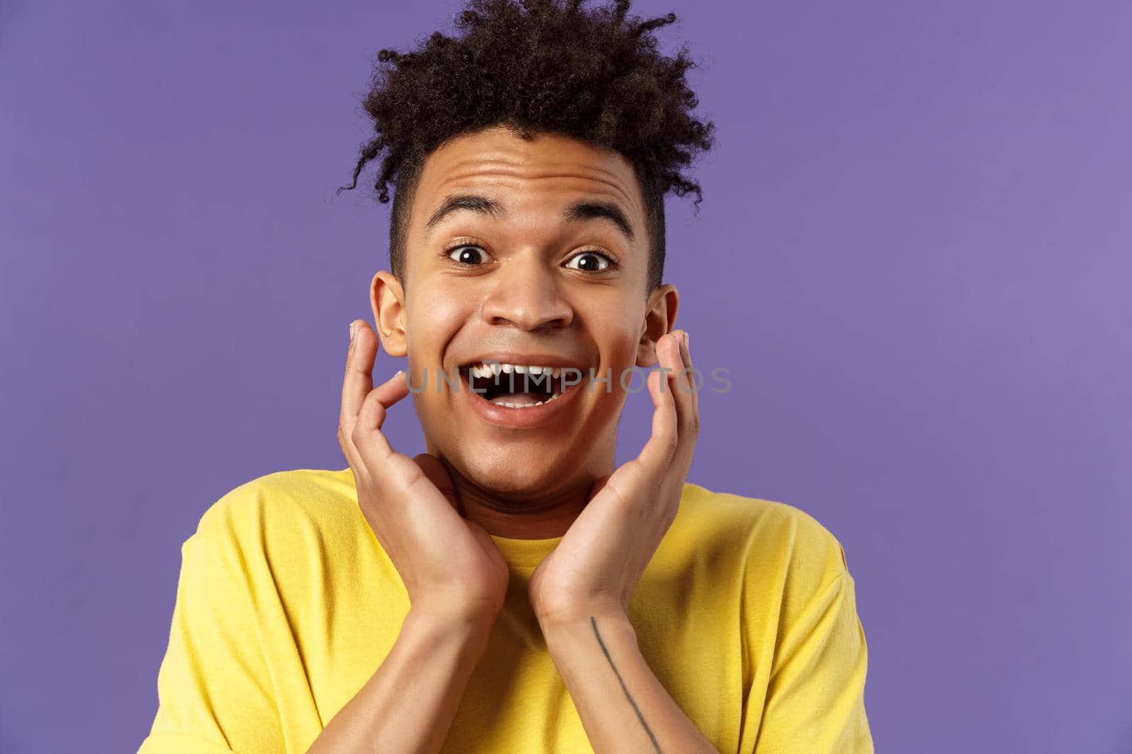 Close-up portrait of extremely happy, enthusiastic young man hear fantastic news, looking surprised and excited, touching face in joy, smiling upbeat look camera astonished, purple background.