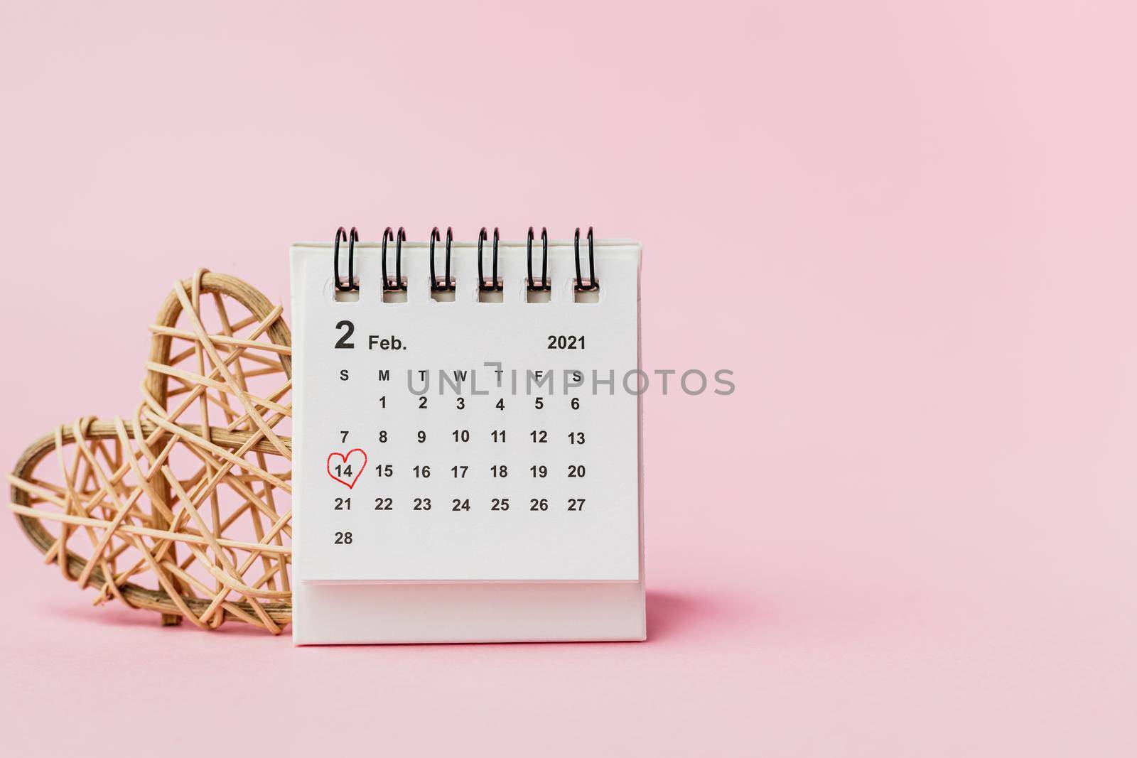 Calendar with red heart shaped marking on date February, 14 and wooden wicker heart against pink background for Valentine's day and love concept