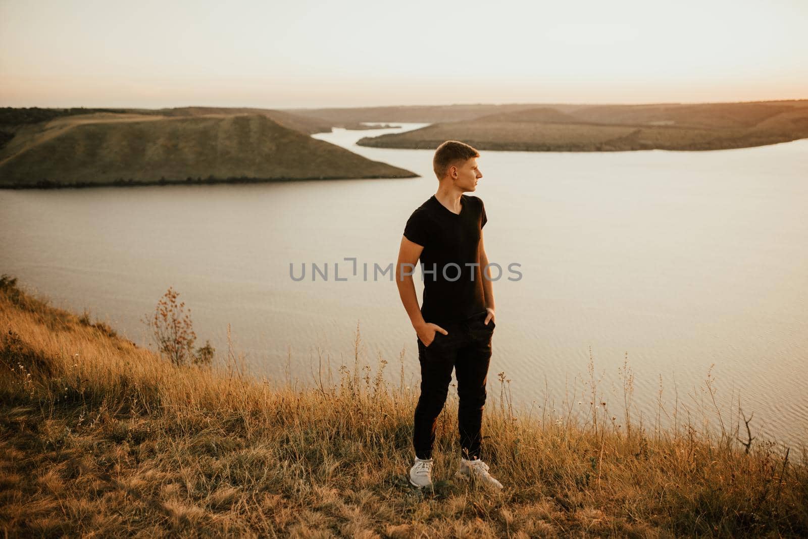Bakota, Ukraine - 08.12.2020: A young athletic man in black casual clothes stands on the side of a mountain against the backdrop of a large lake and islands at sunset.