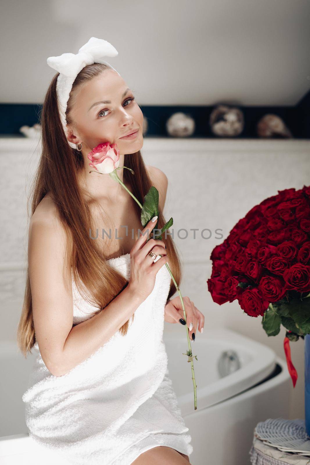 Brunette girl with long hair sitting on bath after morning shower. Pretty lady in white towel looking up and holding flower. Young woman getting bouquet of roses and thinking about boyfriend.