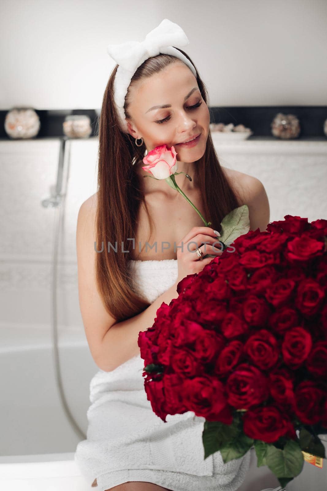 Portrait of gorgeous sensual young brunette woman with headband wrapped in towel sitting in bathroom with bunch of beautiful red roses. She is holding fragile pink rose and smiling at camera. Beauty and skincare concept.