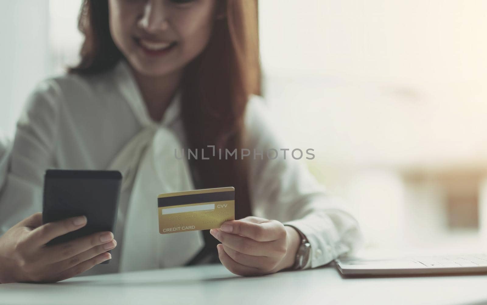 Photo of a happy Asian business woman sitting in the office using a laptop computer and a mobile phone holding a credit card.
