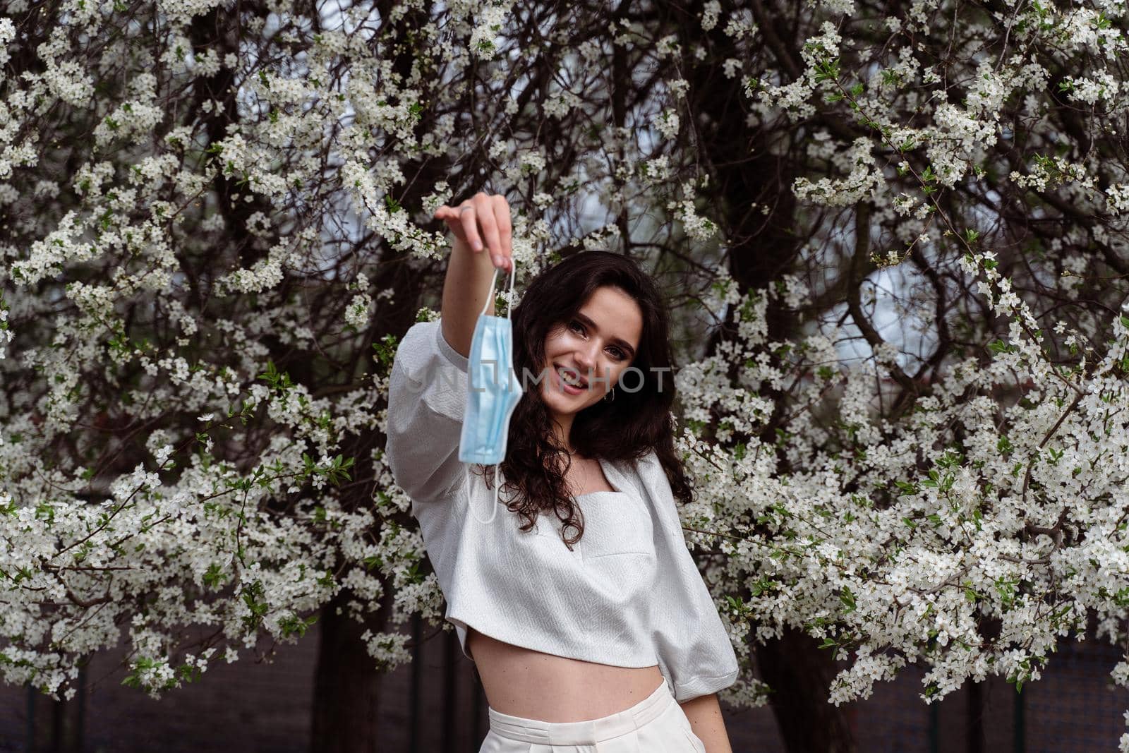 Girl holding medical mask after end of quarantine coronavirus covid-19 period. Model posing near white blooming trees in the garden
