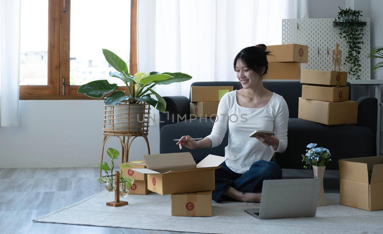 Smiling young Asian business owner woman prepare parcel box and standing check online orders of product for deliver to customer on laptop computer. Shopping Online concept. by wichayada