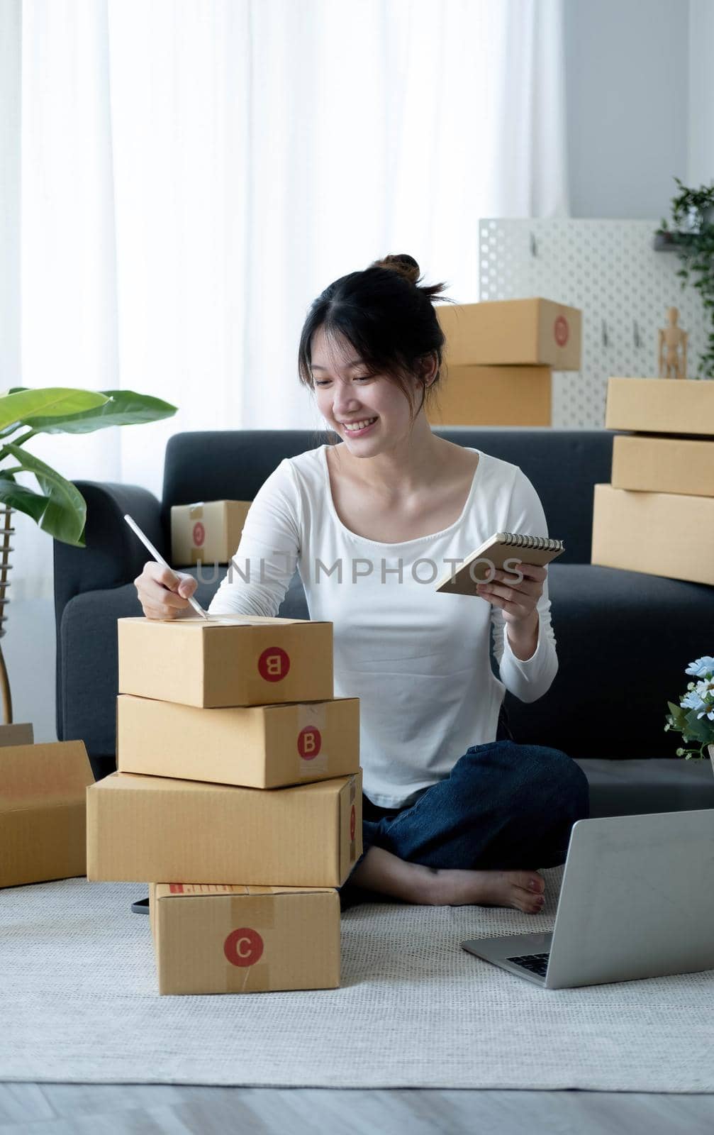 Smiling young Asian business owner woman prepare parcel box and standing check online orders of product for deliver to customer on laptop computer. Shopping Online concept. by wichayada