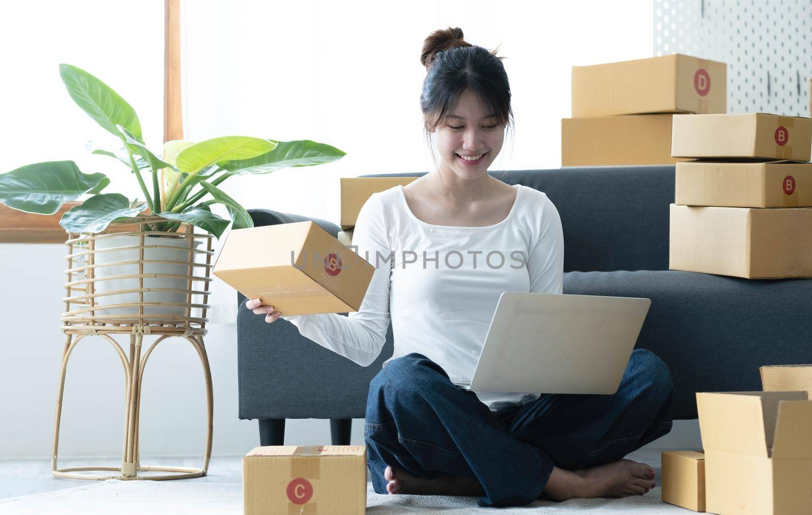 Smiling young Asian business owner woman prepare parcel box and standing check online orders of product for deliver to customer on laptop computer. Shopping Online concept..