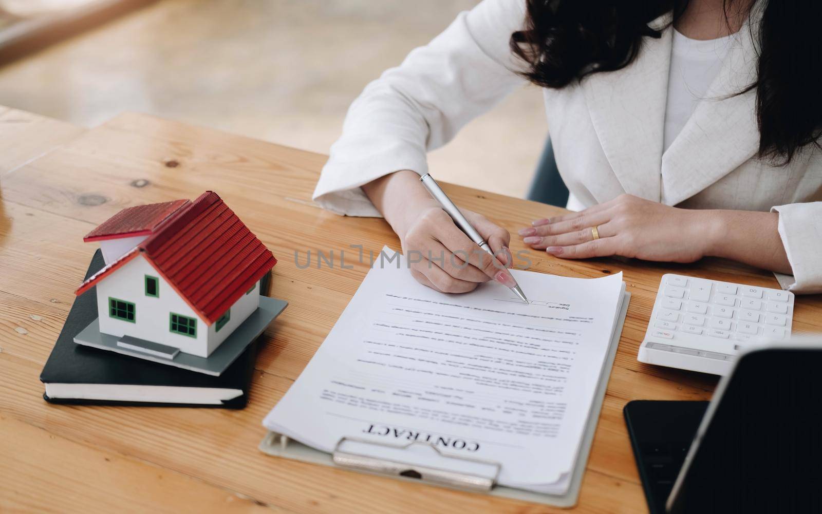 Cropped image of real estate agent assisting client to sign contract paper at desk with house model by wichayada