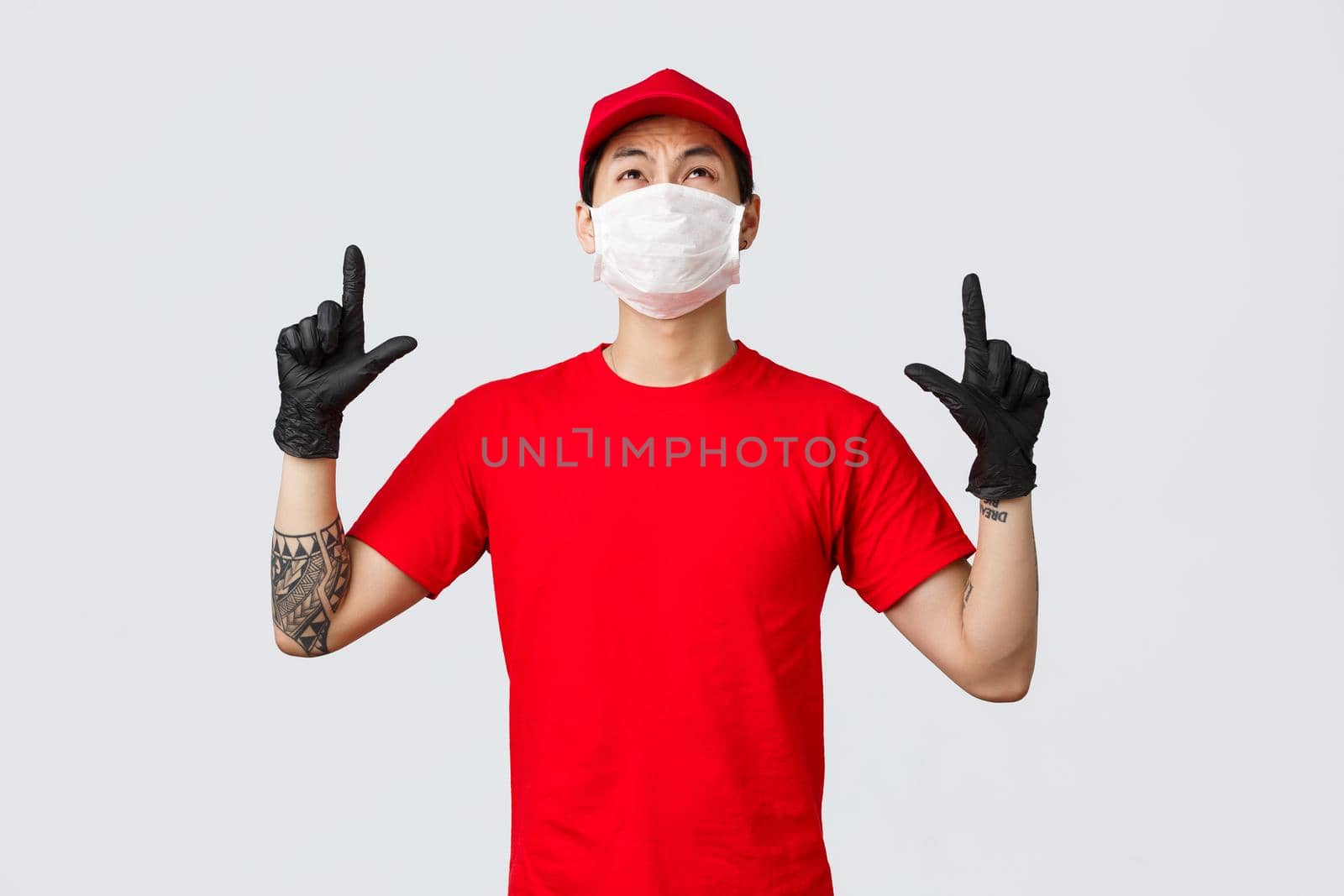 Disappointed and reluctant asian courier in red t-shirt and cap, grimacing from dislike and aversion as looking pointing up, showing unpleasant thing on top, wear uniform while deliver goods.