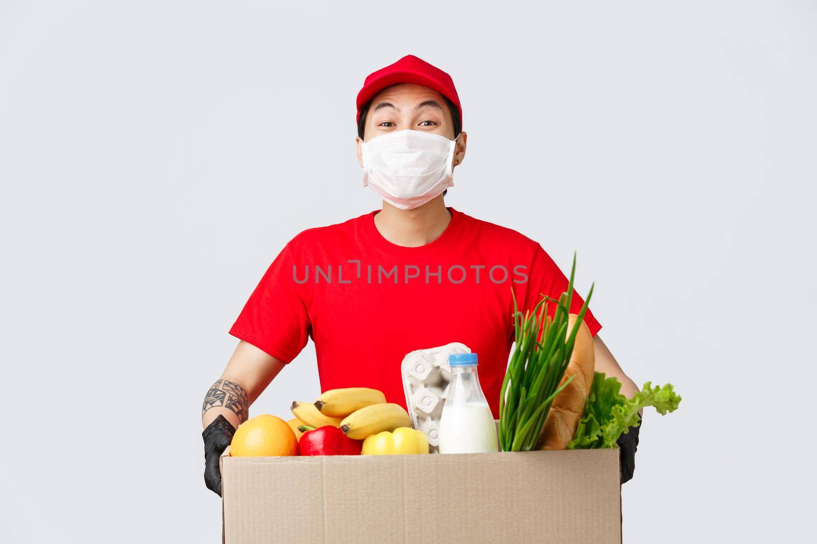 Online shopping, food delivery and coronavirus pandemic concept. Smiling delivery man in red uniform, holding box with fresh groceries, wear medical mask and gloves, contactless shopping.
