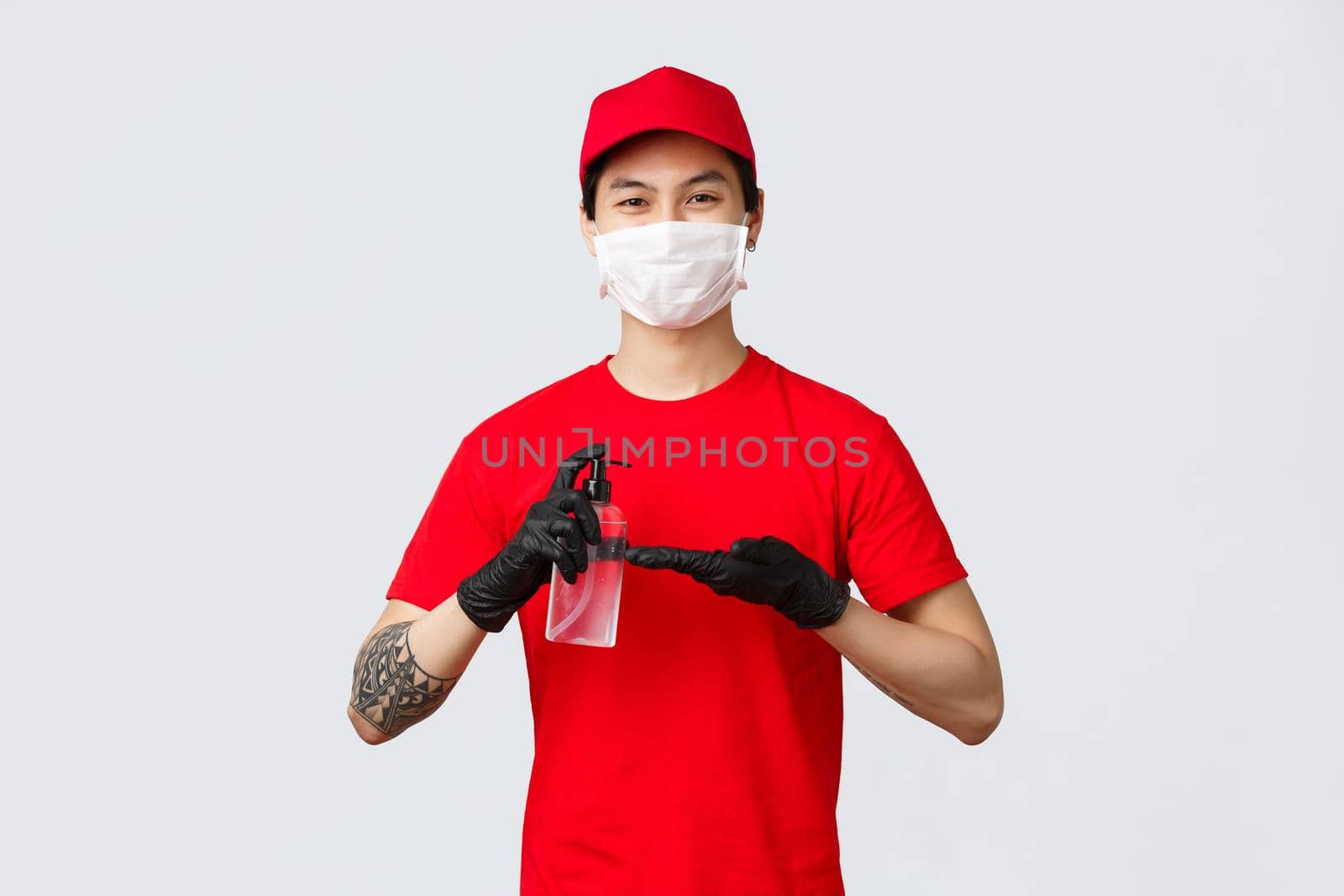 Smiling delivery man in red cap and t-shirt, wear medical mask and gloves, apply hand sanitizer to protect from covid-19, virus during outbreak. Courier cares about clients, use sanitizer during work.