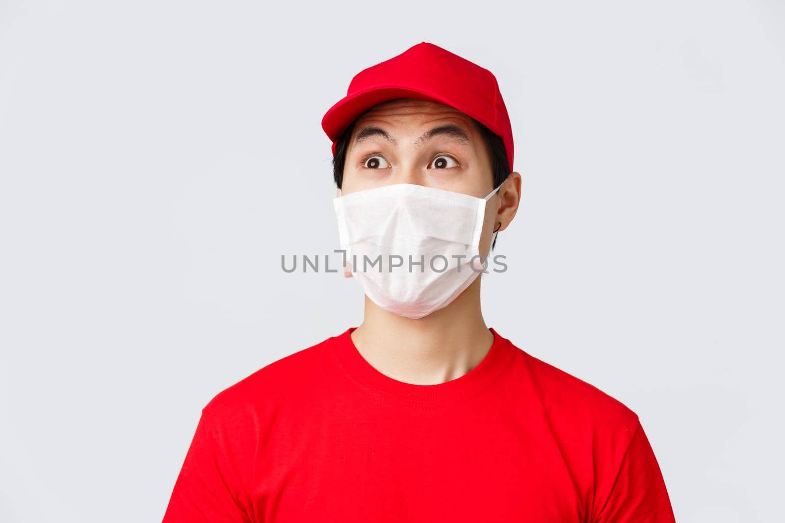 Covid-19, self-quarantine online shopping and delivery concept. Excited and impressed courier in medical mask and uniform red cap, t-shirt, staring upper left corner astonished, grey background.