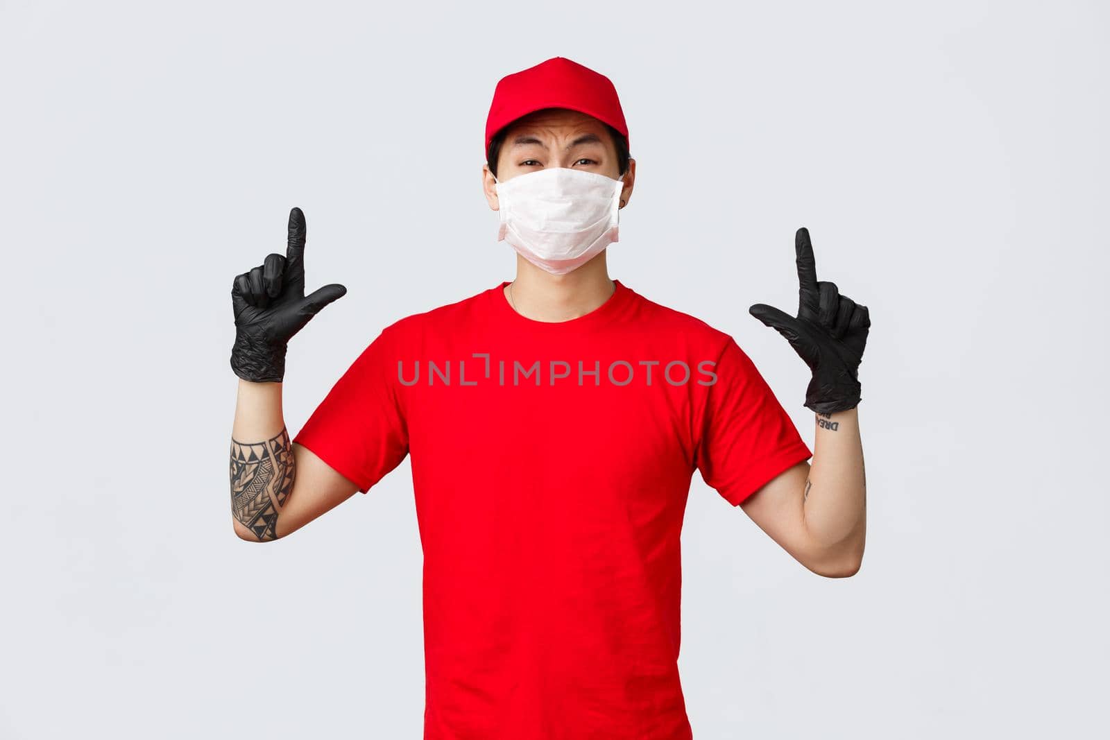 Reluctant or scared, insecure asian guy whining, grimacing as pointing fingers up, wear delivery uniform, red cap and t-shirt, company protect personnel health with protective masks and gloves.