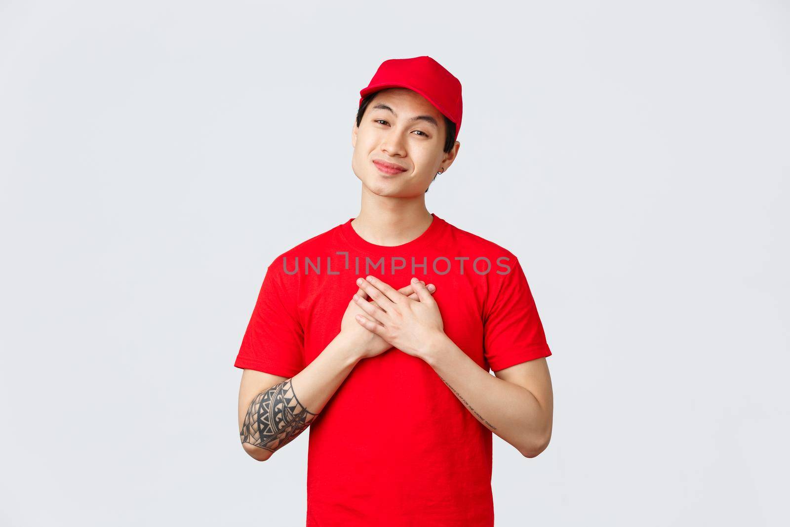 Express delivery, shipping and logistics concept. Touched happy asian delivery guy in red cap and t-shirt, hold hands on heart, sighing and gazing at lovely warm gesture of kindness.