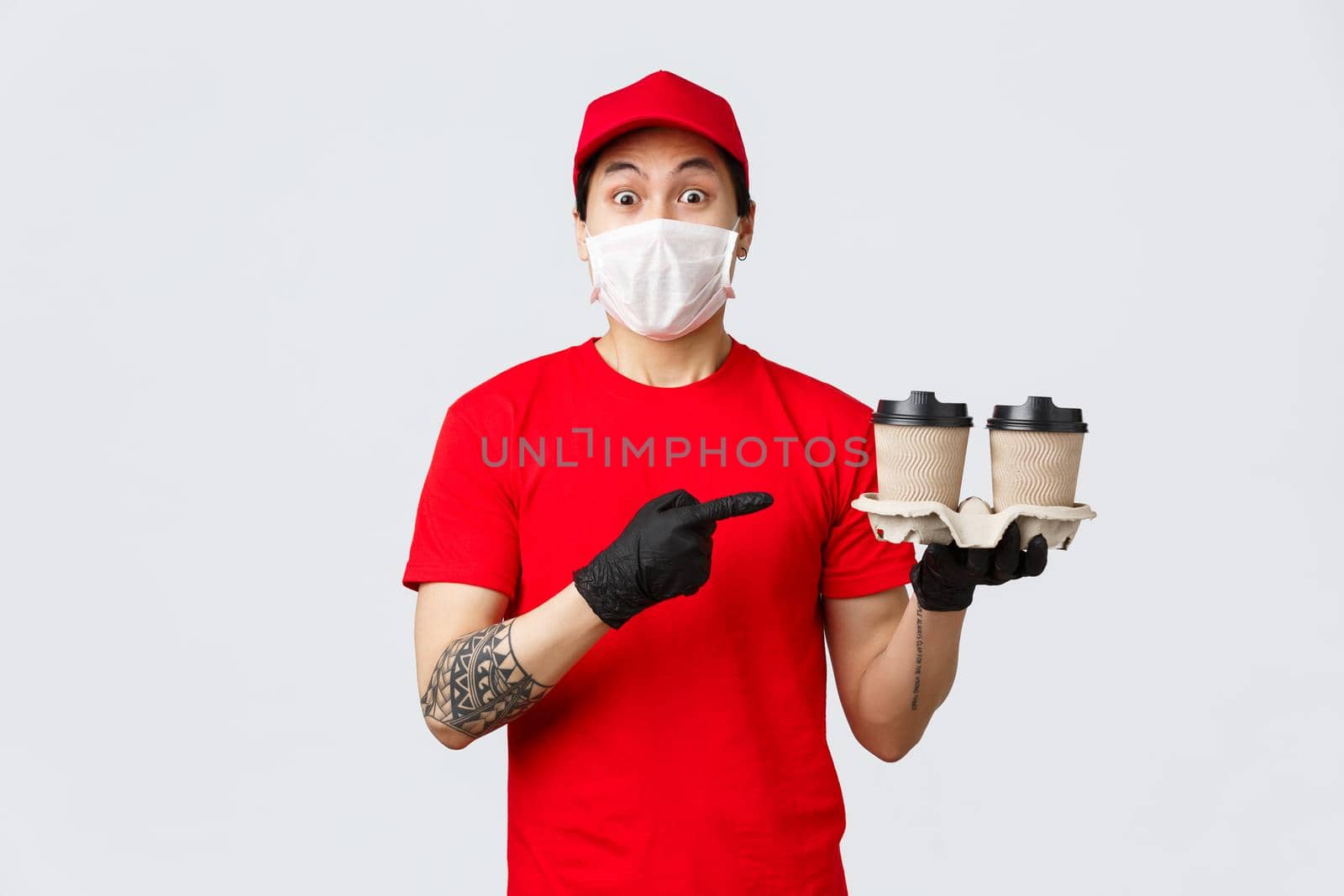 Excited asian delivery man in red cap, medical mask and gloves, safely deliver your beverege, holding cups of beverage, pointing at coffee and staring amazed camera, courier working during covid 19.