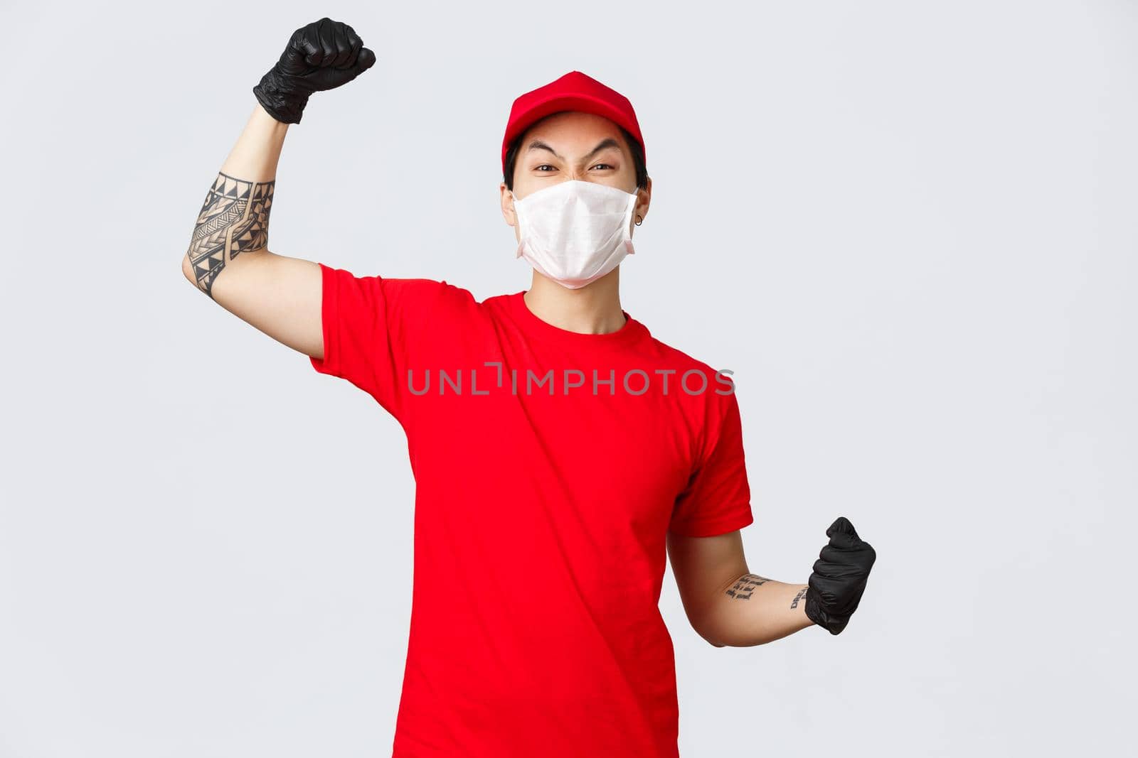 Cheerful enthusiastic delivery guy in red t-shirt and cap, wearing medical mask, protective gloves, raising hand up chanting, say yes or hooray, celebrate success, encourage order online.