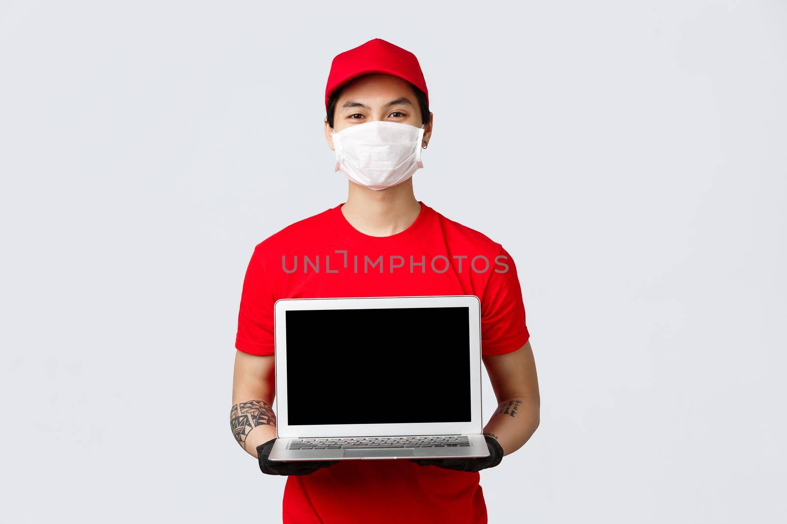Place your order and we deliver fast. Smiling asian delivery guy showing online shopping site for clients during pandemic covid-19 wear protective medical mask and gloves, holding laptop.