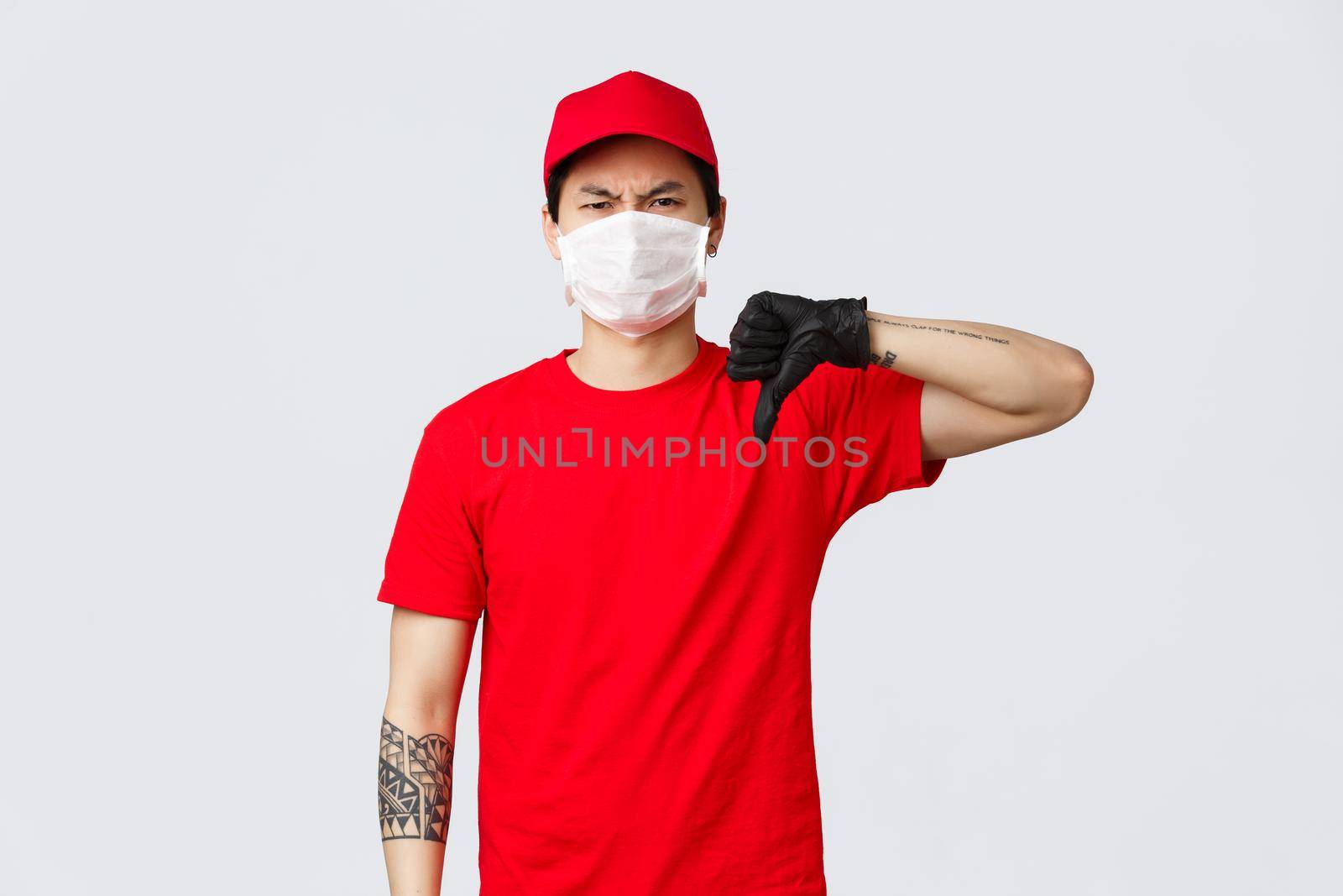 Another deliveries awful. Reluctant, disappointed delivery man in red cap, t-shirt, wearing protective gloves and medical mask, grimacing dislike and disagree, show thumb-down negative bad opinion.