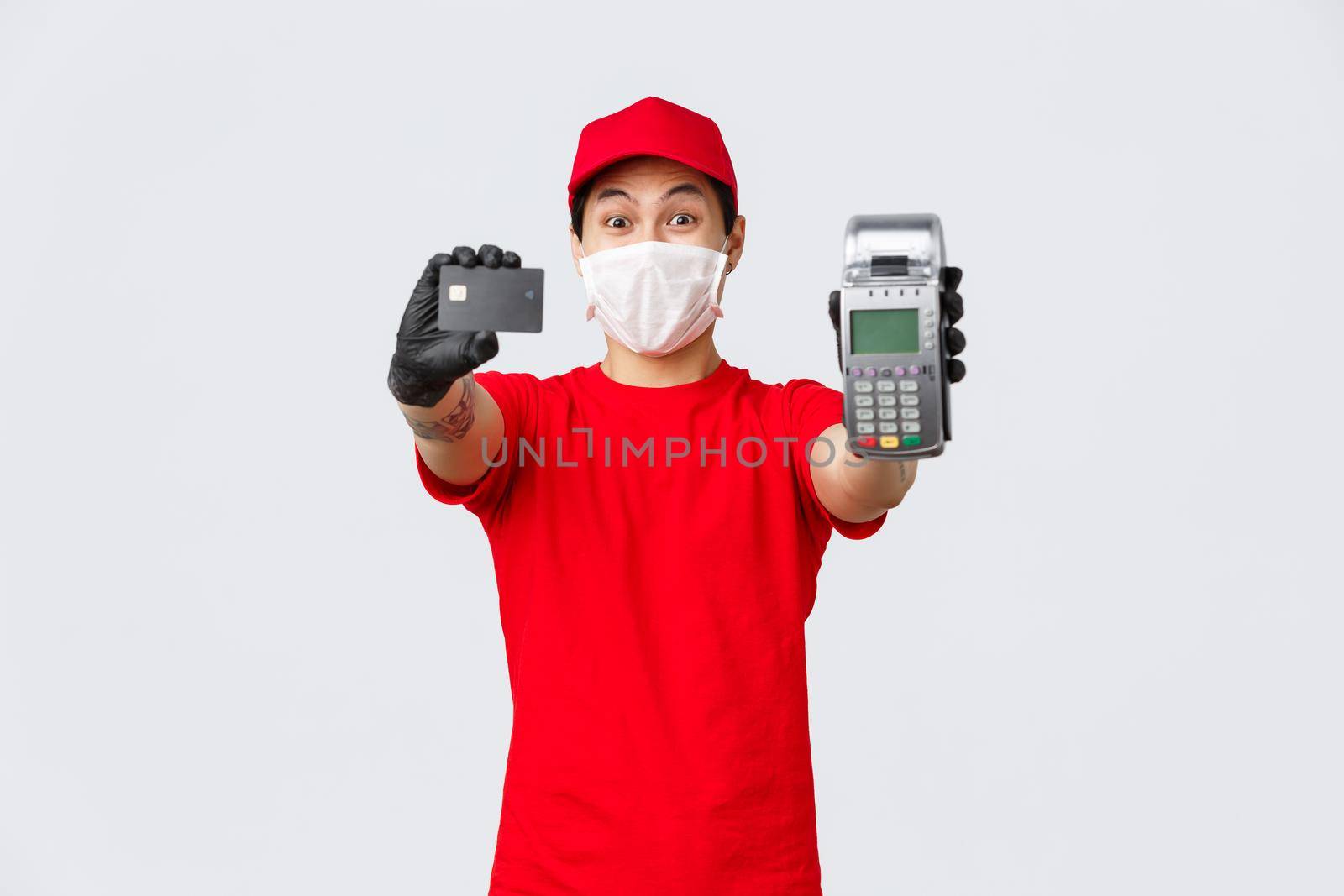 Contactless delivery, safe purchase and shopping during coronavirus concept. Excited asian courier in red uniform, medical mask and protective gloves recommend use credit card and POS terminal.
