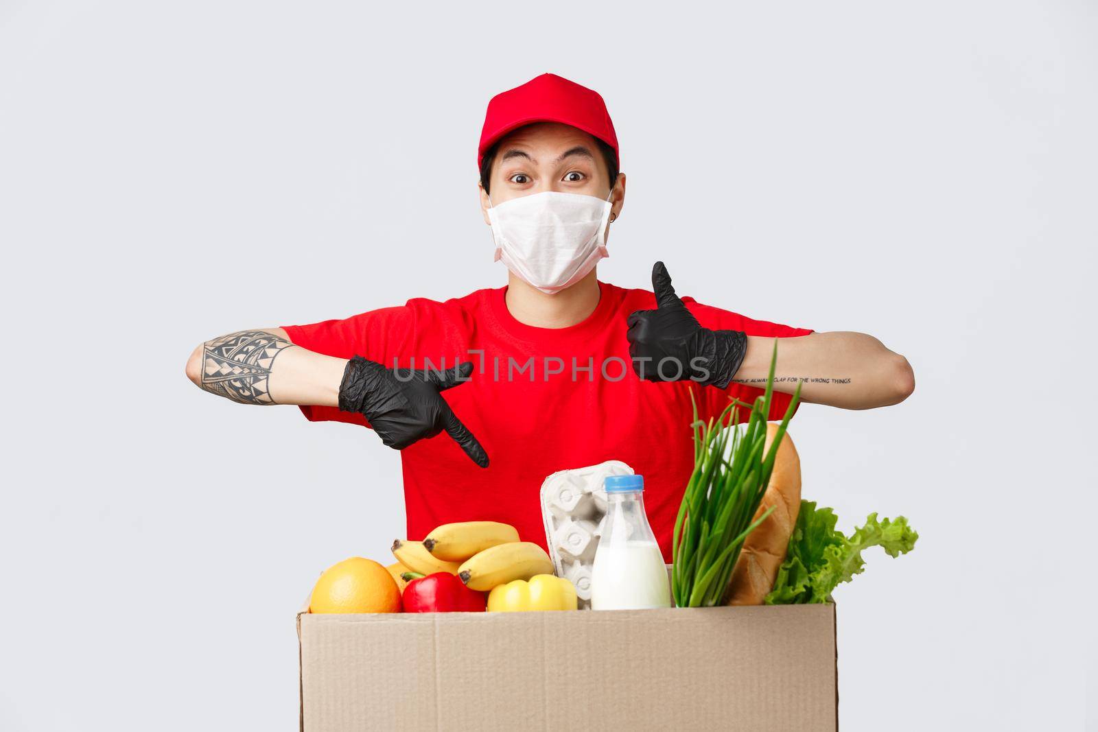 Online shopping, food delivery and coronavirus pandemic concept. Charismatic delivery guy in red uniform, medical mask and gloves, show thumb-up and approval, pointing at grocery parcel.