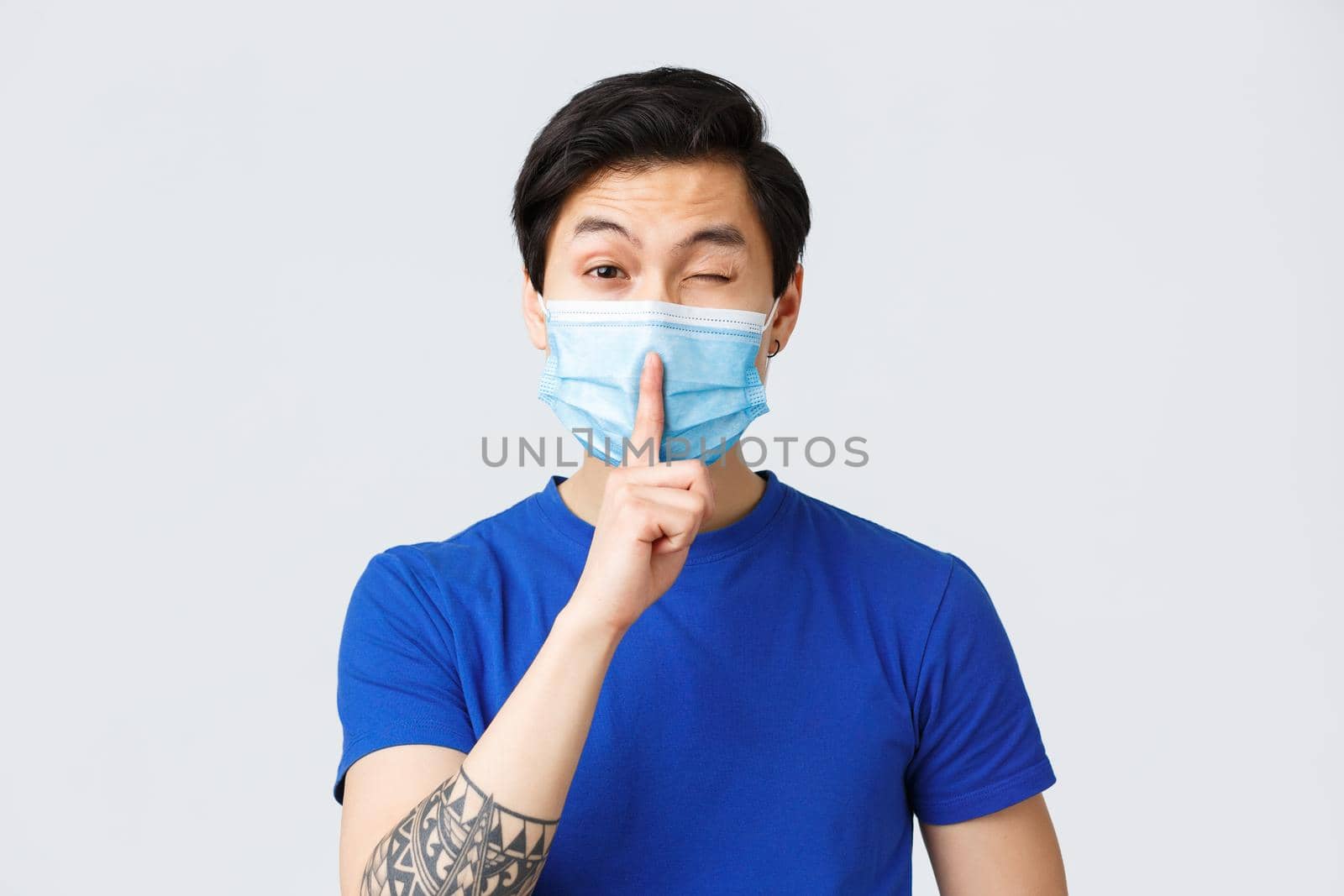 Different emotions, lifestyle and leisure during coronavirus, covid-19 concept. Cheeky handsome asian man in medical mask promise keep secret, shush and wink coquettish, be silent.