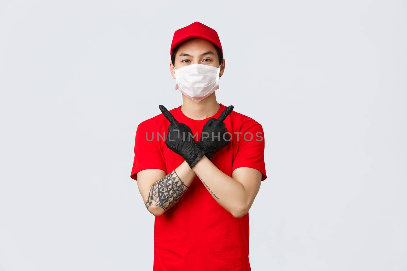Young confident delivery guy in red t-shirt and cap, wear protective gloves and medical mask to deliver parcels at customer home during covid-19, showing variants, pointing sideways, two choices.