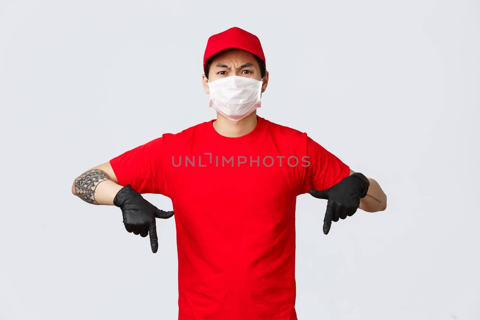 Angry young asian delivery guy complaining over rude customer, pointing fingers down in protective gloves and medical mask, frowning upset, stare disappointed at camera, wear courier uniform.