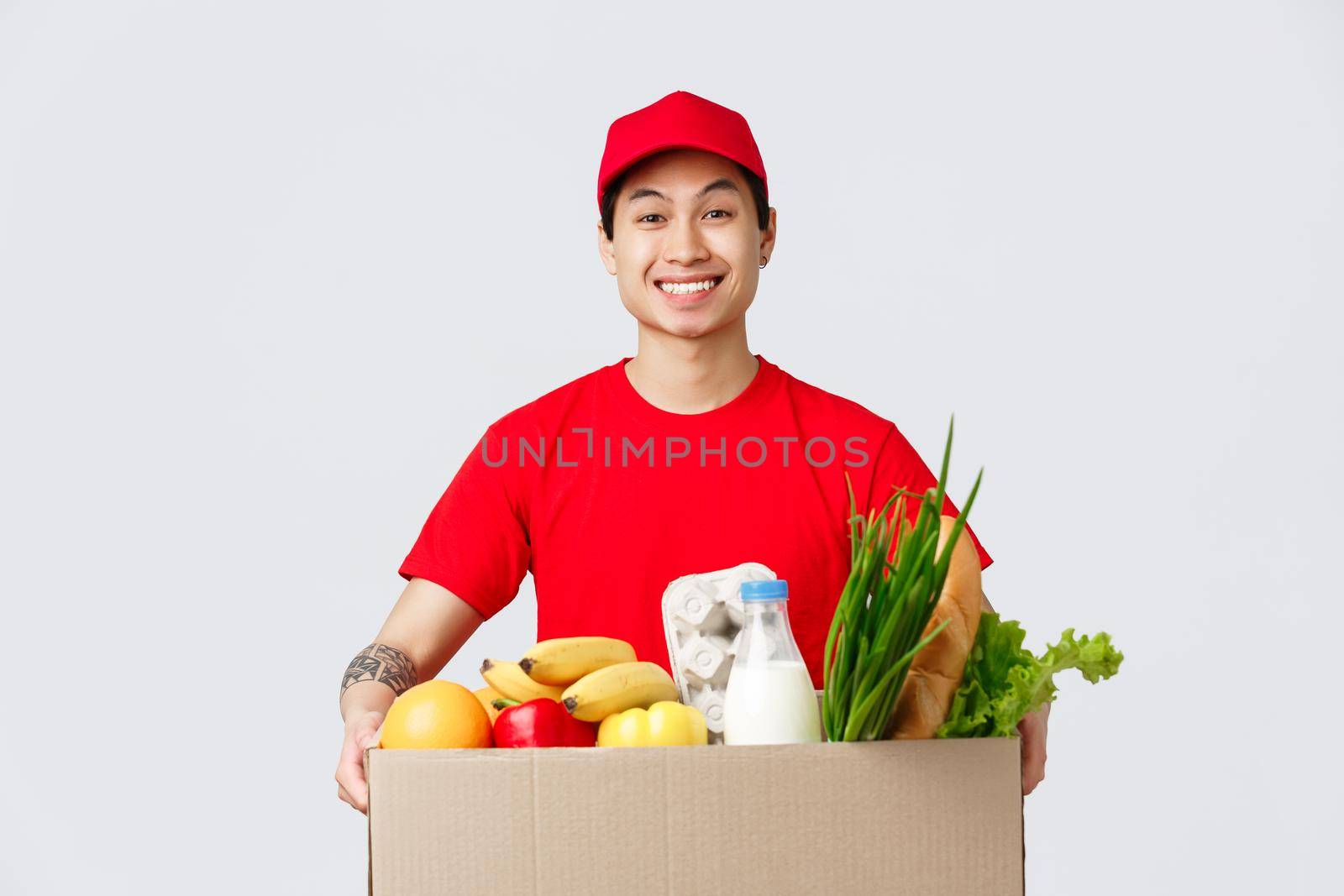 Online shopping, food delivery and internet stores concept. Cheerful smiling asian guy in red cap and t-shirt, holding box with fresh groceries, standing customer door, employee deliver product.