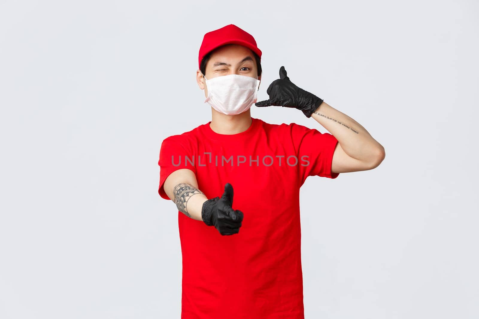 Call out customer support anytime. Cheerful asian delivery guy in medical mask and protective gloves, wink and show thumb-up, phone sign, provide safe courier service. carrier and shopping concept.
