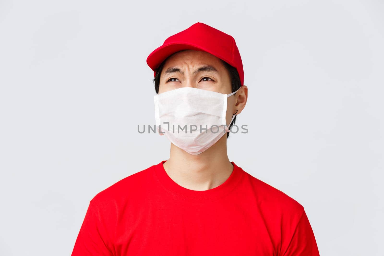 Covid-19, self-quarantine online shopping and delivery concept. Complaining reluctant courier in red t-shirt and cap, medical mask, whining about night shifts at work, look disappointed.