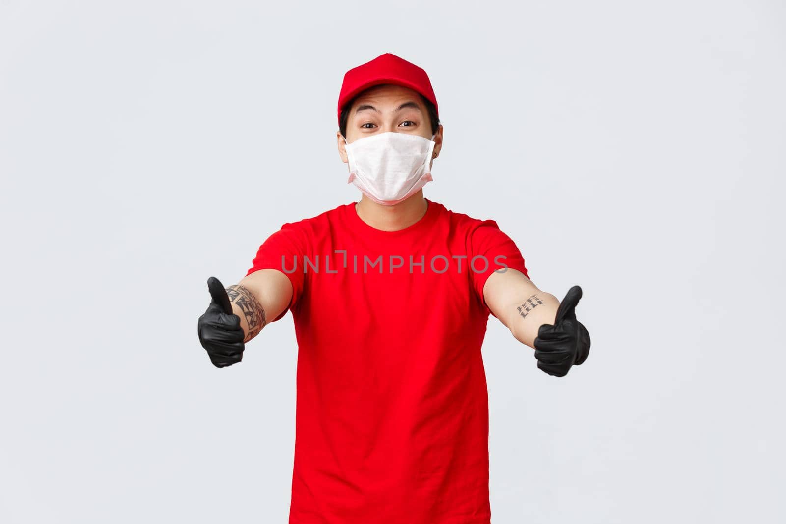 Cheerful smiling asian courier in red cap and t-shirt stretch hands forward to take parcel from client at delivery company. Deliver guy in uniform, medical mask and gloves receive shipping goods.