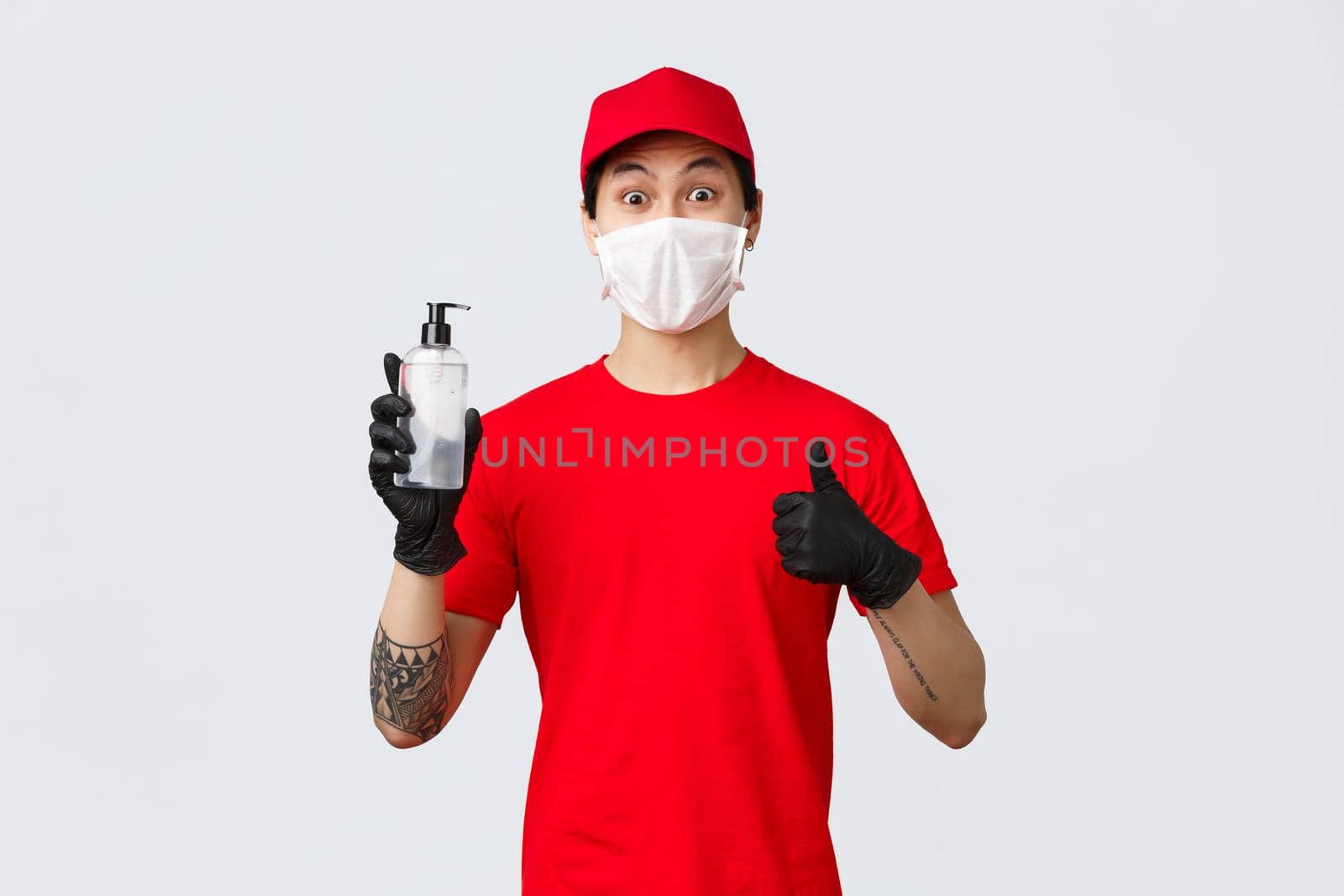 Amused delivery man in red cap and t-shirt, make thumb-up sign, wear protective gloves and medical mask, provide clients and company workers with hand sanitizers. Concept of contactless safe delivery.