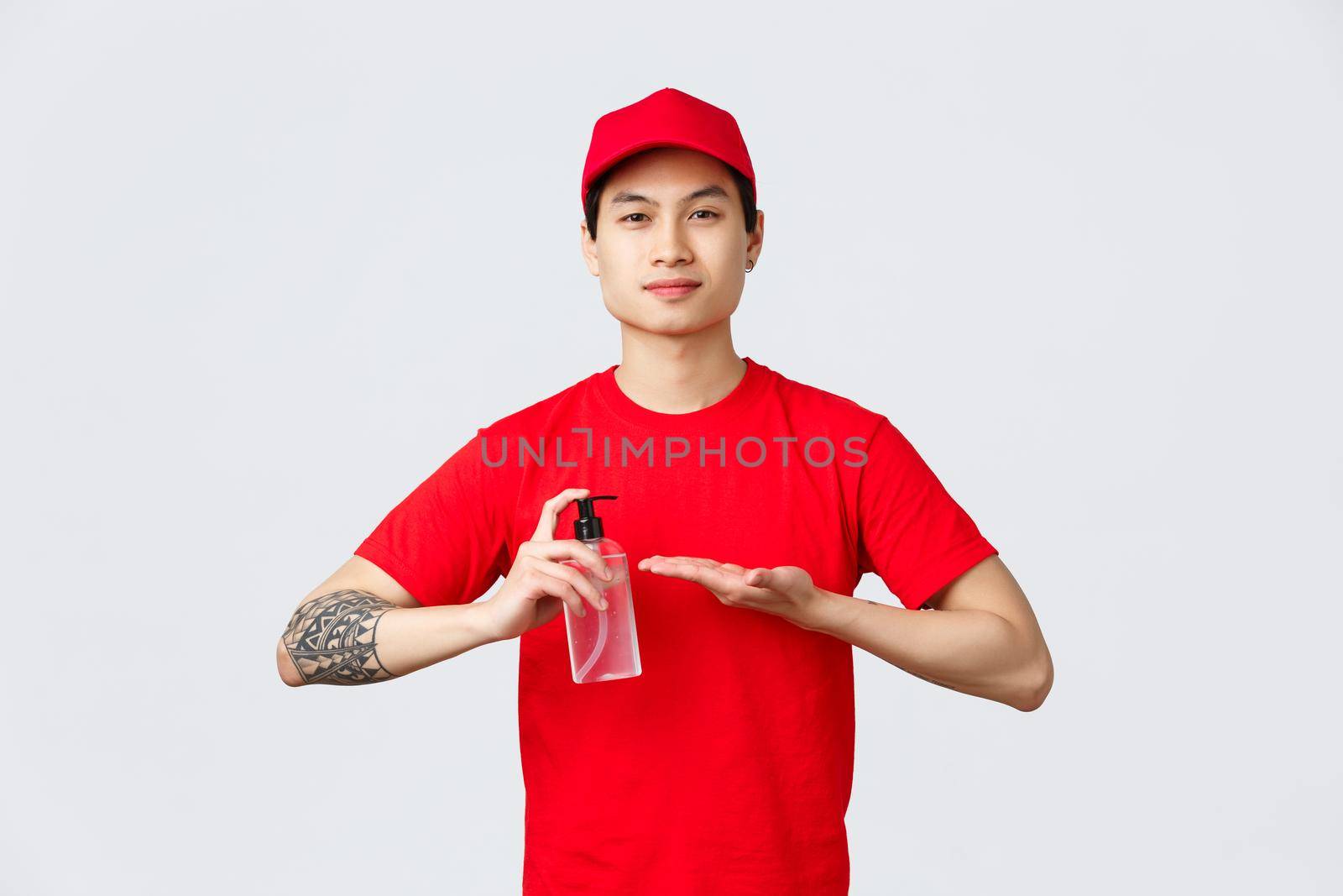 Quarantine, safe contactless delivery and online shopping concept. Smiling asian delivery guy in red cap and t-shirt, apply hand sanitizer on hands to prevent spread of covid-19 and viruses.