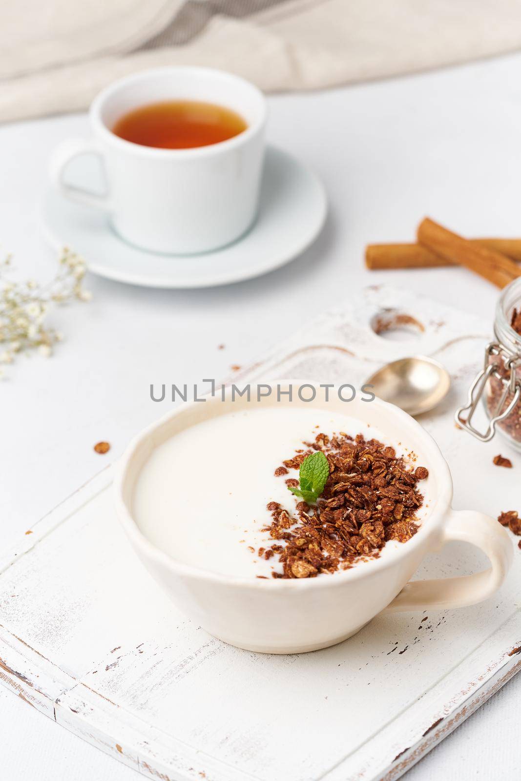 Yogurt with chocolate granola in cup, breakfast with tea on white wooden background, vertical