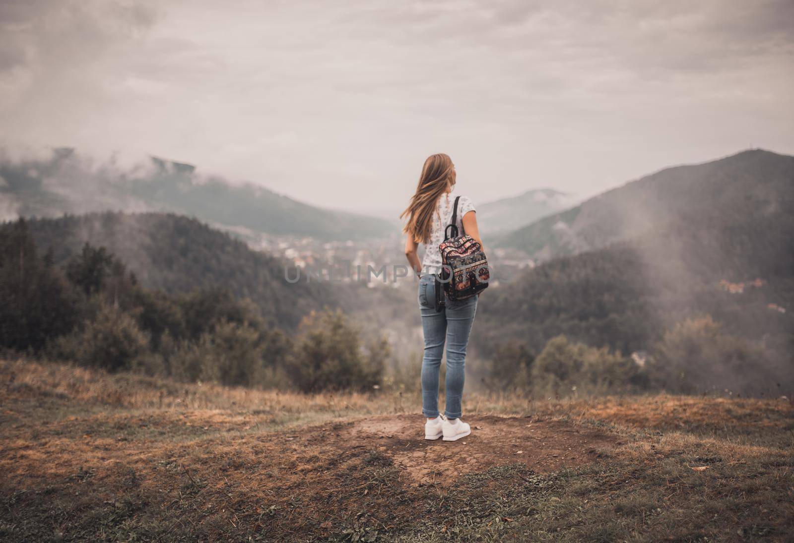 Young blonde woman standing alone with backpack on wild forest mountains at background. European girl wear tight jeans and white t-shirt sneackers. Carpathian Mountain peaks in fog scenery landscape