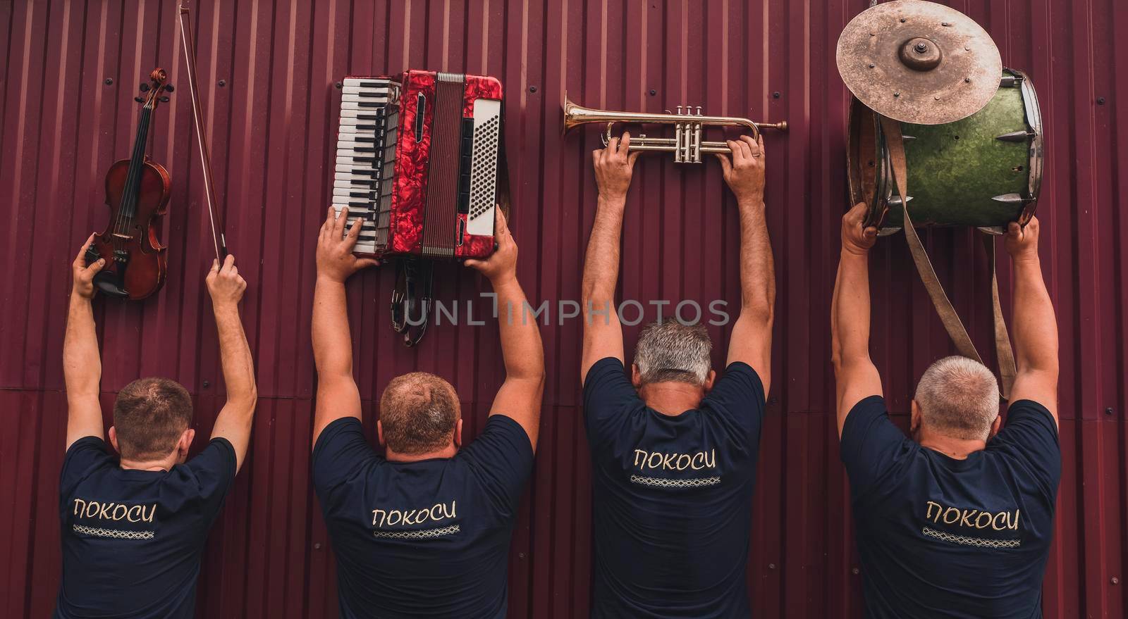 Inscription:haymakings men. men stand with backs and raised their hands up above their heads holding their musical instruments:violin,accordion,trumpet,drumms.musical group.Red high metal fence