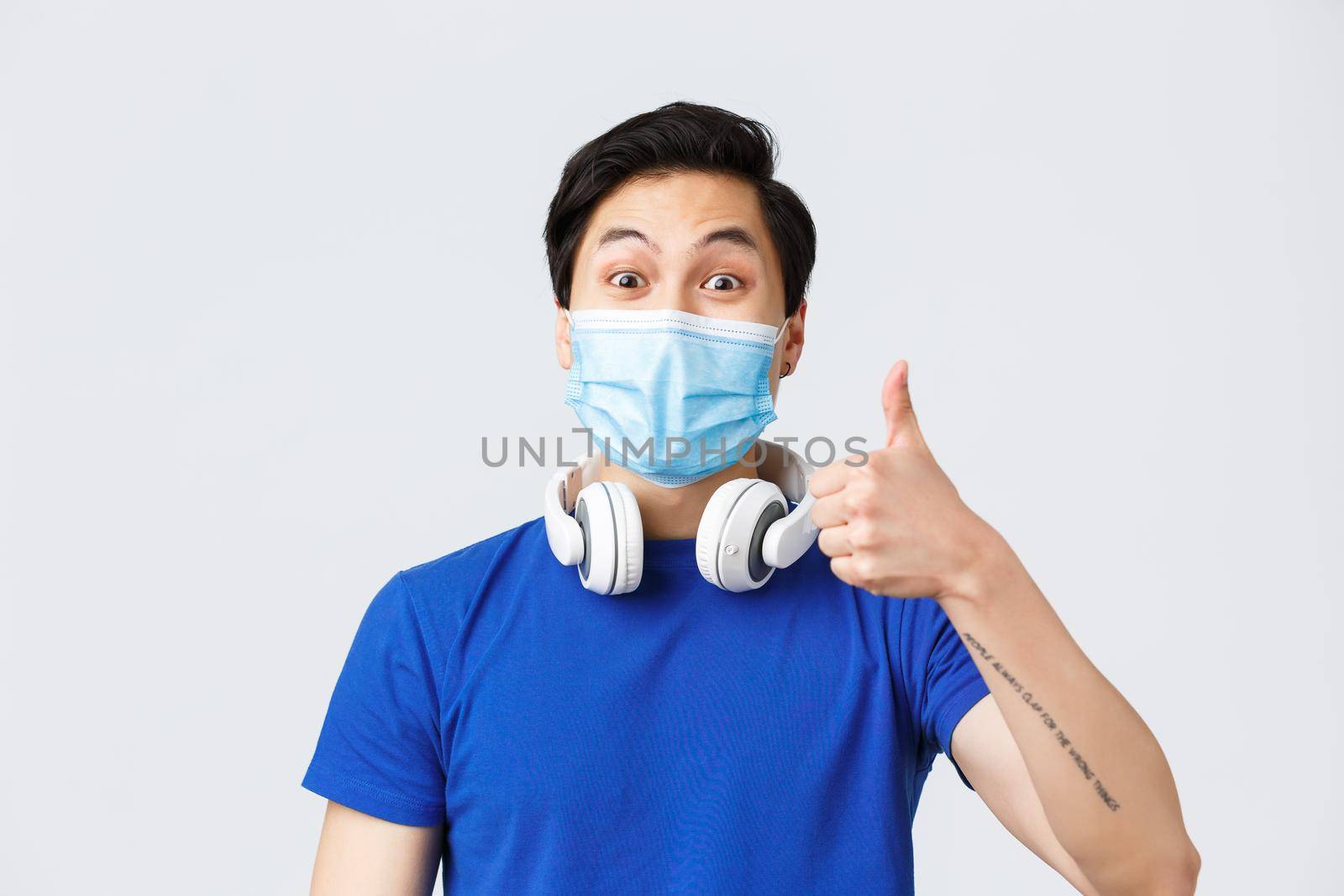 Different emotions, lifestyle and leisure during coronavirus, covid-19 concept. Close-up of upbeat asian guy in medical mask show thumb-up in approval of awesome new song playing in headphones.