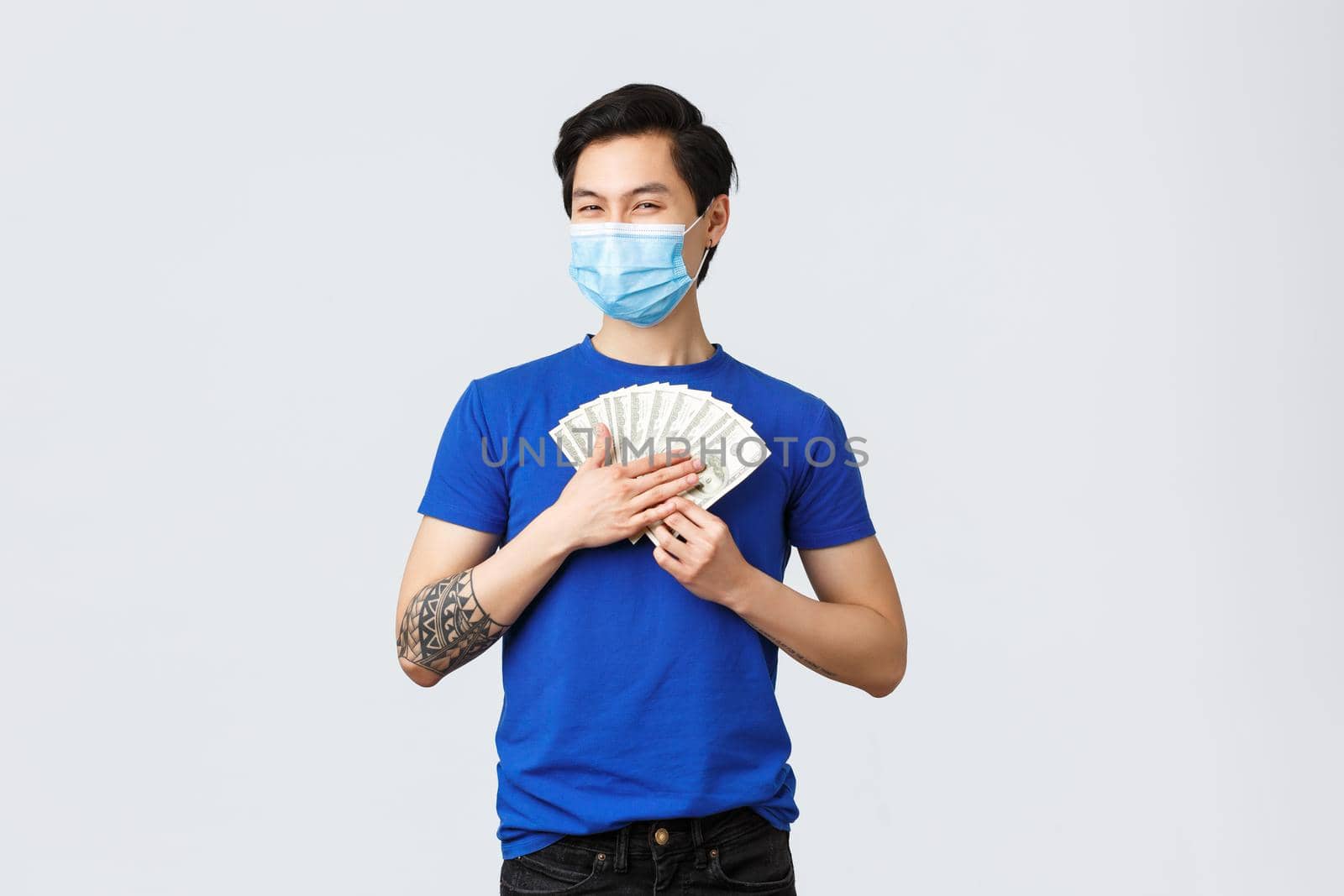 Money, lifestyle, insurance and investment concept. Satisfied griedy asian guy hugging cash of dollars and smiling pleased, wearing medical mask, earned or won lottery.