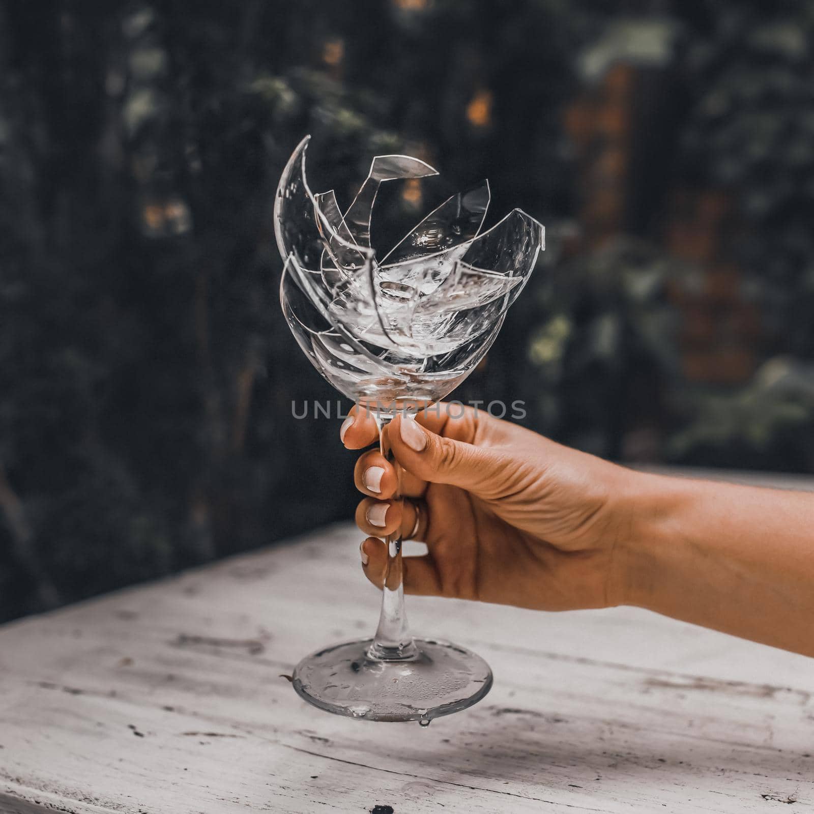 Broken crystal goblet in the hand of a woman man. Against the background of a light gray wooden shabby table and a green leafy wall. Shards folded into a glass. Ring in a glass.