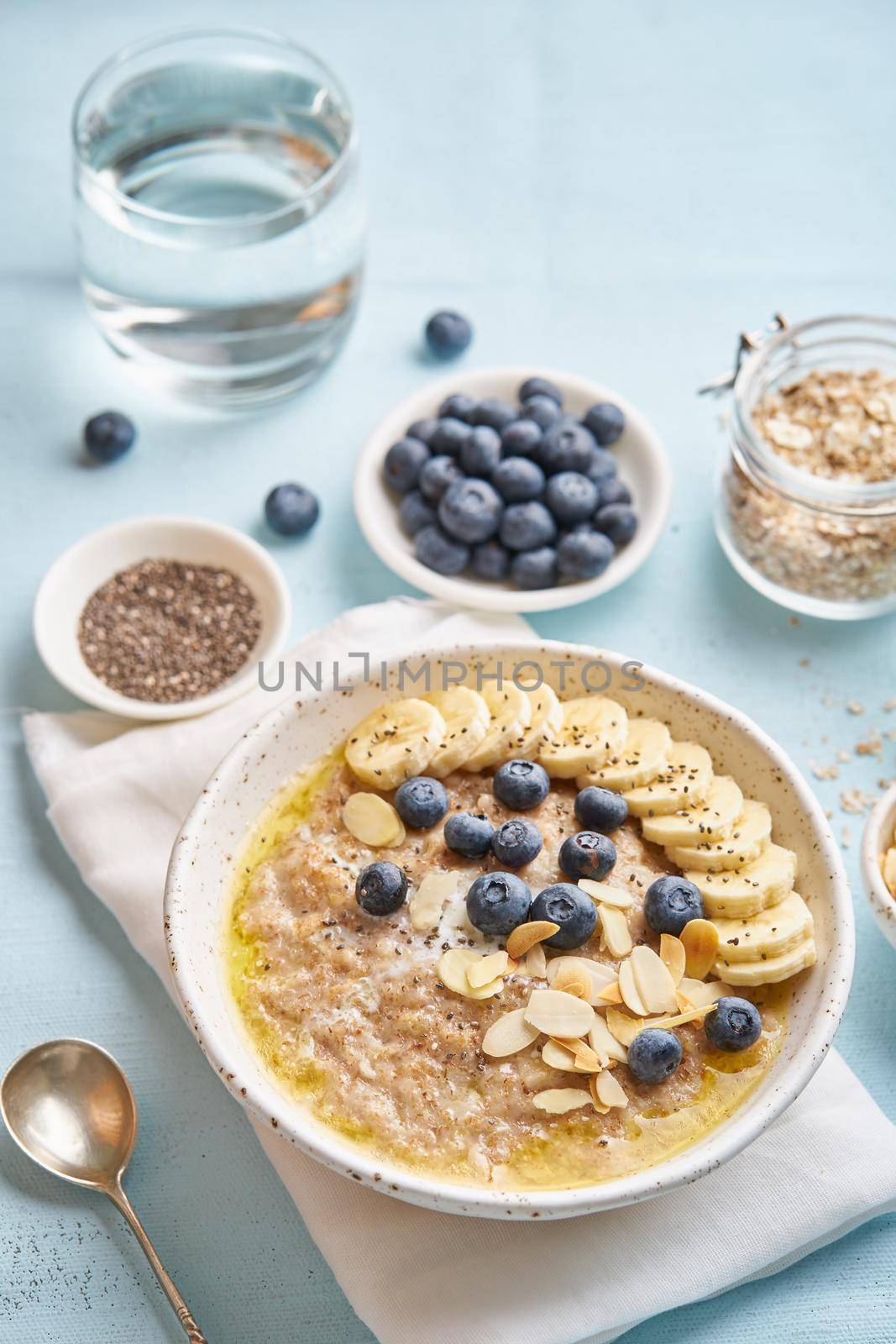 Oatmeal with blueberries, banana and cup of water on blue light background. Side view, vertical. Healthy diet breakfast