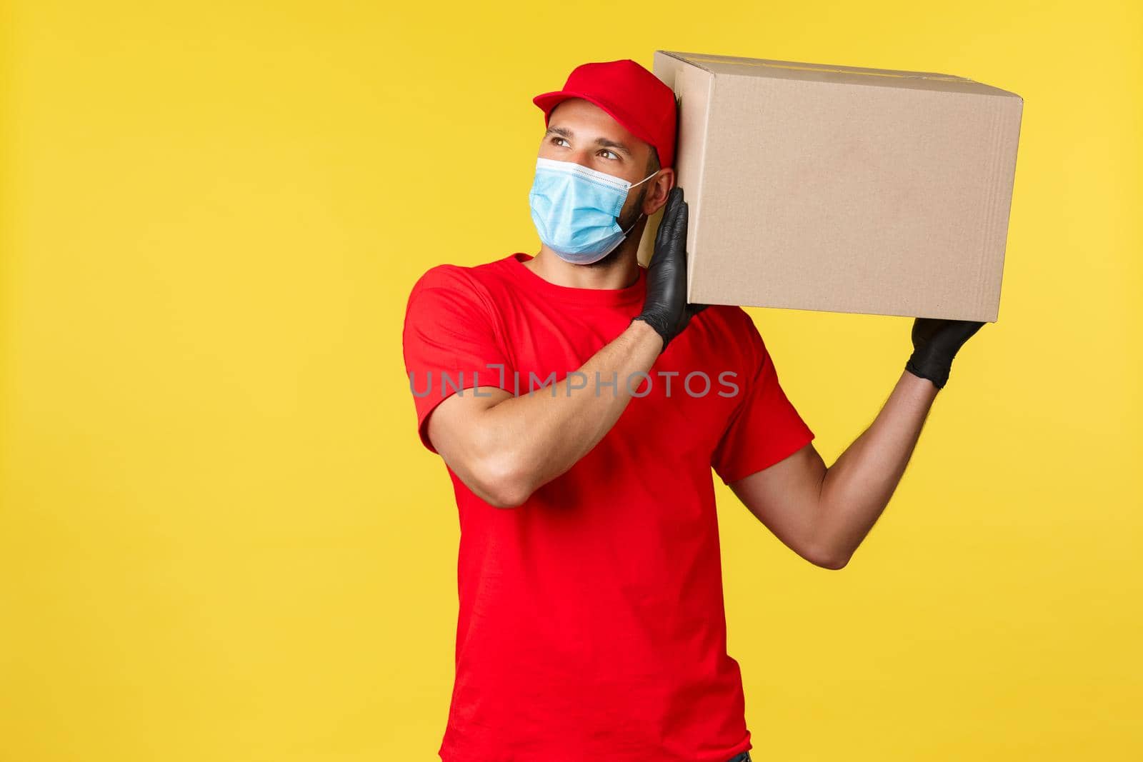 Express delivery during pandemic, covid-19, safe shipping, online shopping concept. Young courier protect clients order, carefully holding package on shoulder, look upper left corner, medical mask.