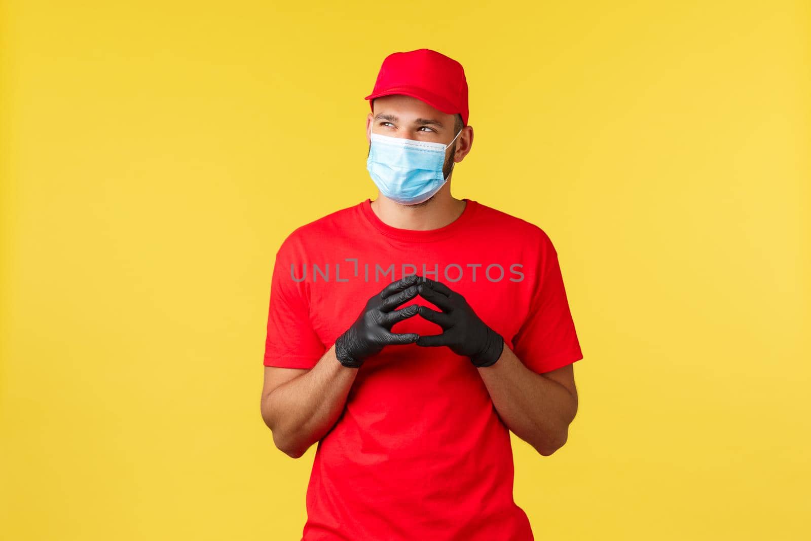 Express delivery during pandemic, covid-19, safe shipping, online shopping concept. Sly and thoughtful courier in red uniform, gloves and medical mask, thinking, steeple fingers, look away.