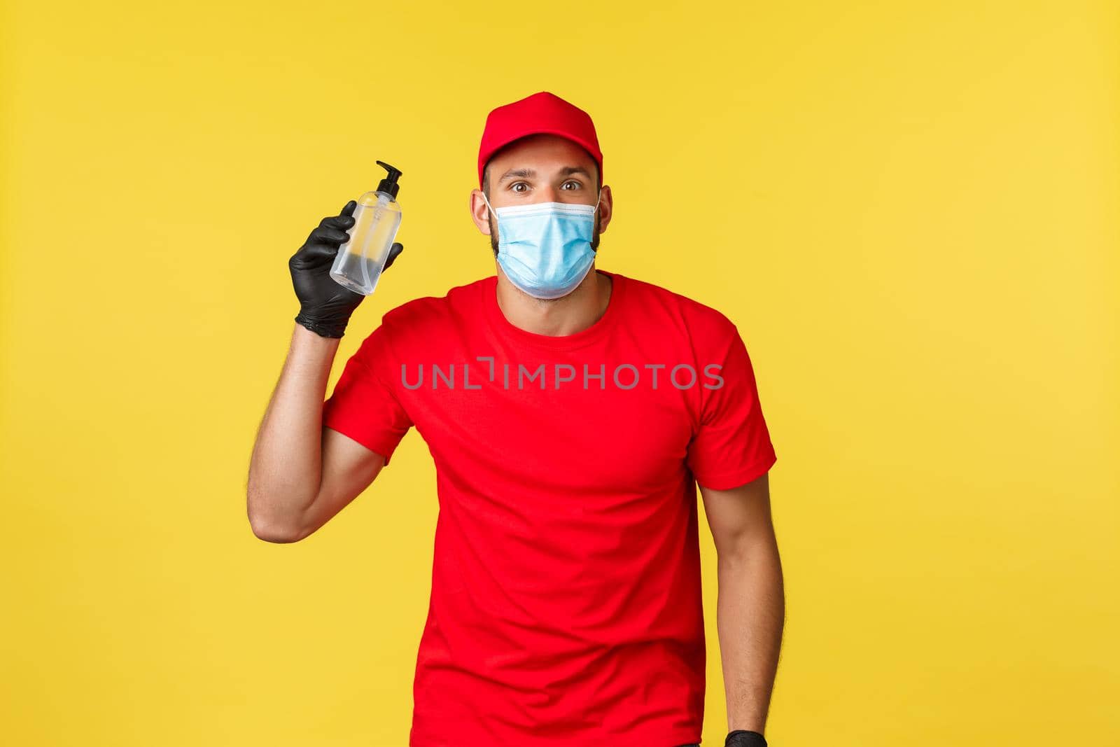 Covid-19, delivery orders, shopping, contactless handling and social distancing concept. Cheerful courier in red uniform, medical mask and gloves, recommend new hand sanitizer.