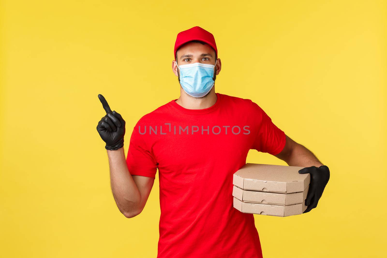 Food delivery, tracking orders, covid-19 and self-quarantine concept. Courier in red uniform and medical mask, provide contactless fast deliver, pointing finger left, holding pizza.