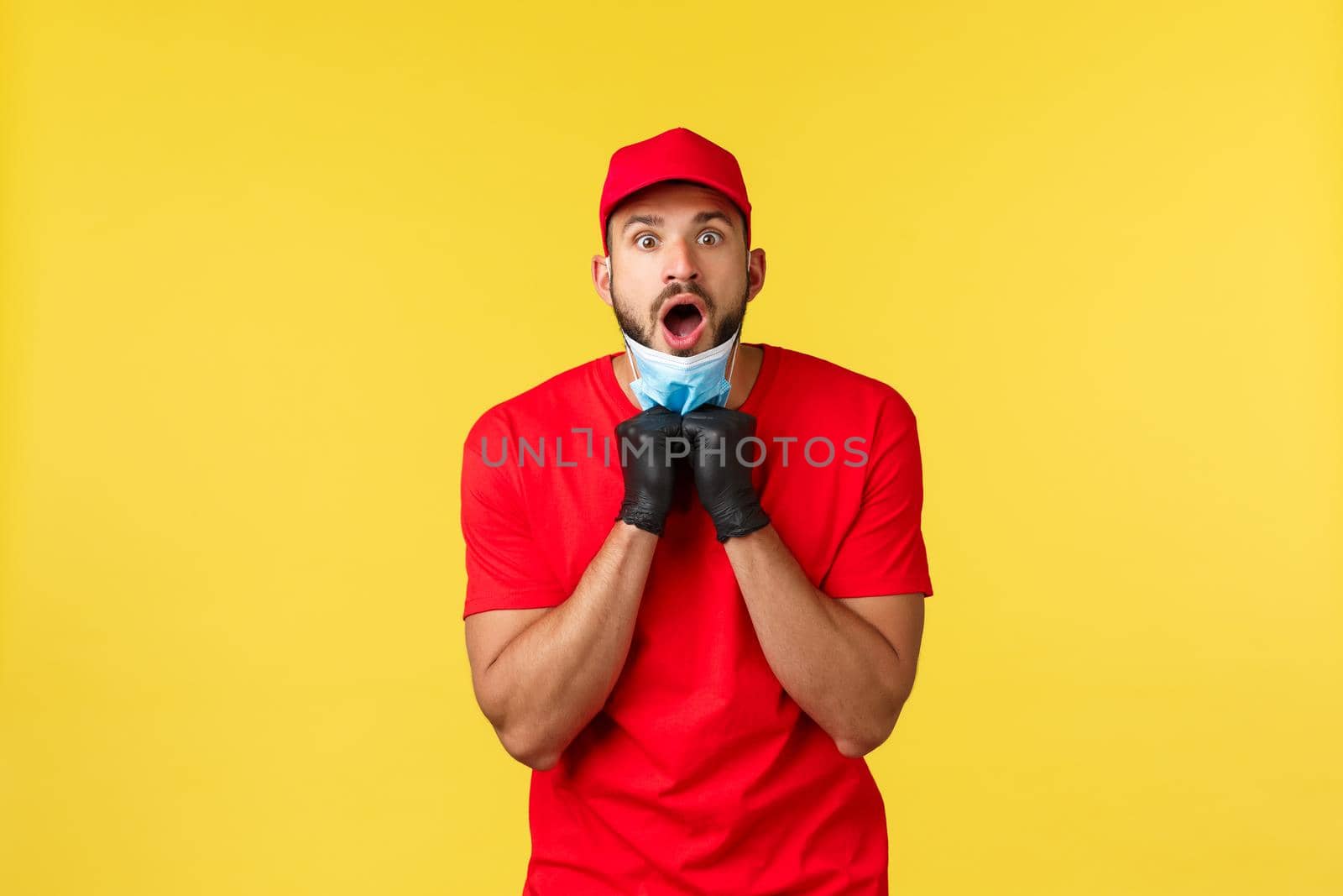Express delivery during pandemic, covid-19, safe shipping, online shopping concept. Shocked and startled courier in red uniform, gloves, take-off medical mask and open mouth saying wow.