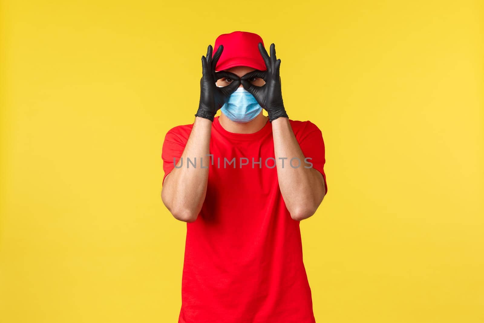 Express delivery during pandemic, covid-19, safe shipping, online shopping concept. Funny courier in red uniform, medical mask and gloves, show okays or glasses with hands.