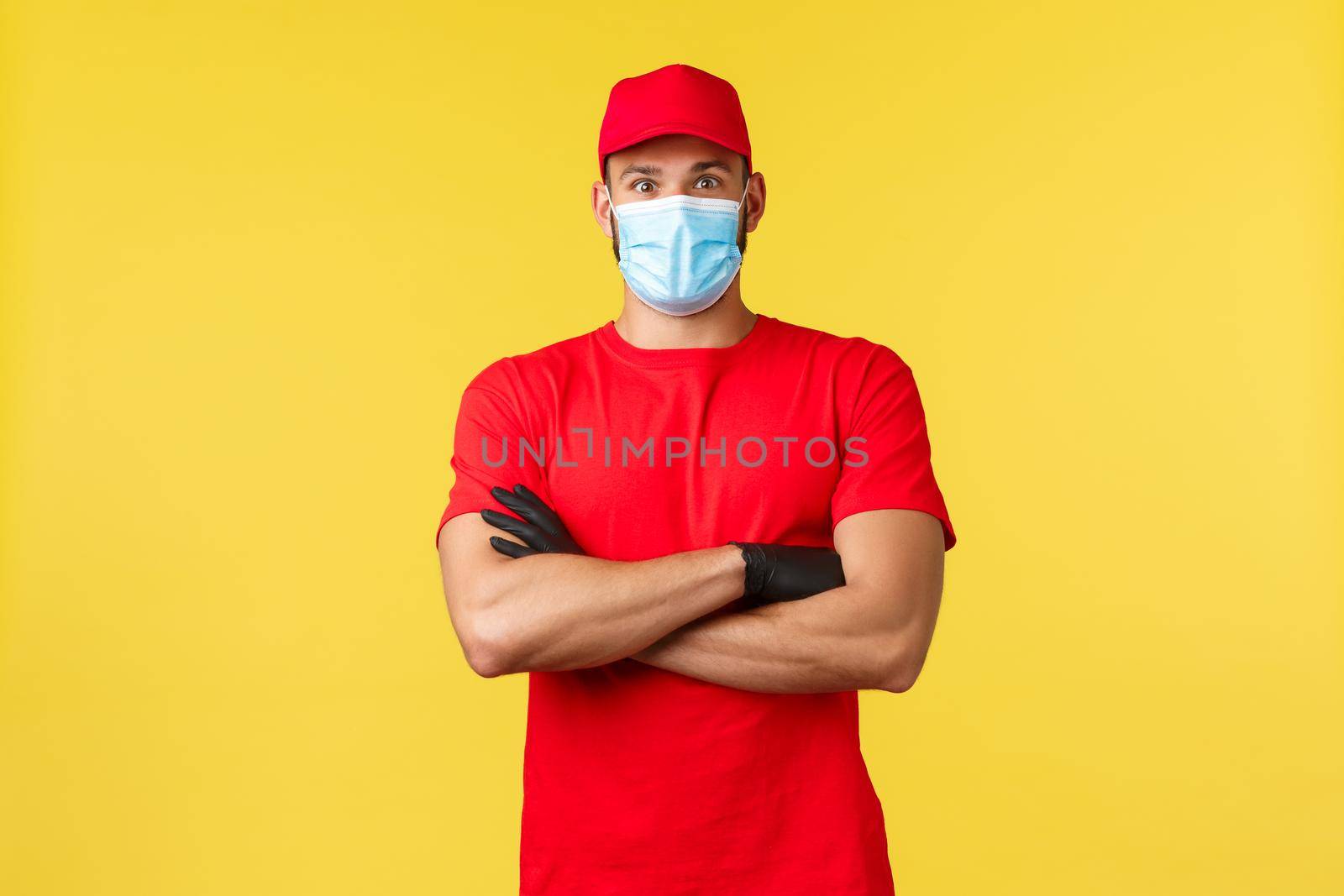 Express delivery during pandemic, covid-19, safe shipping, online shopping concept. Surprised male courier in red uniform cap and t-shirt, cross arms chest, medical mask, stand yellow background.